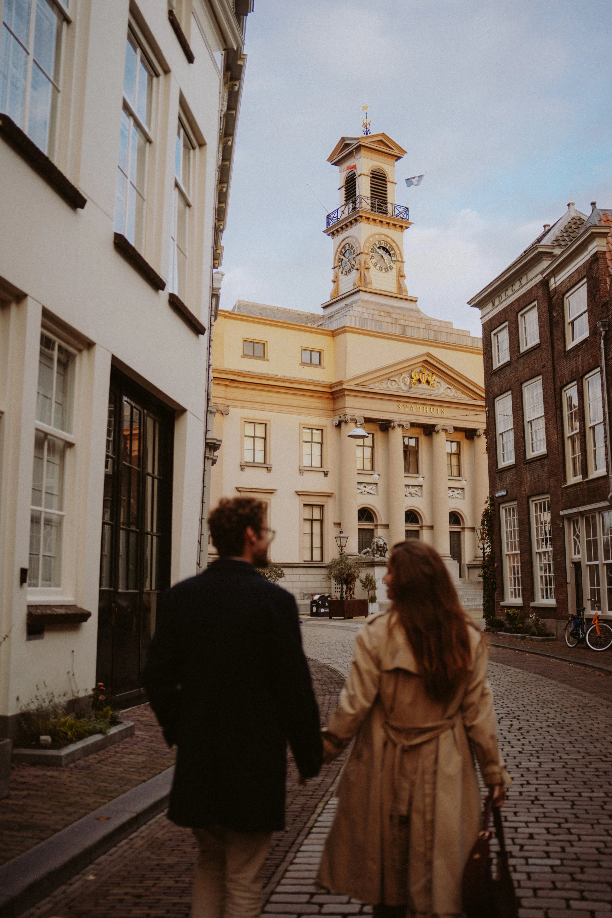 A man and woman holding hands walking on cobblestone street in front of a classical building with a clock tower and the word "Stadhuis" on it, in a European city during evening.