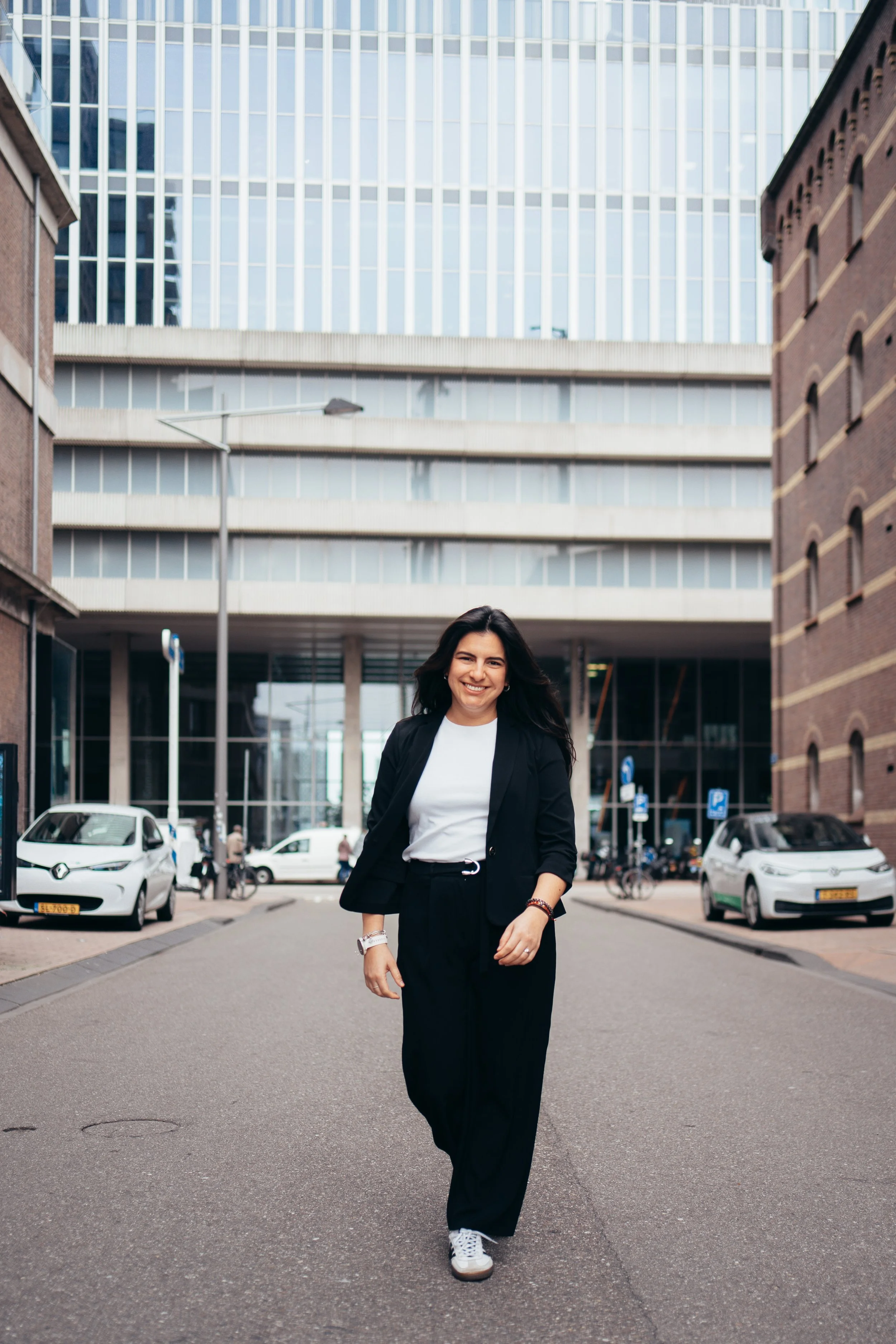 Woman with dark hair and a smile walking on a city street, wearing a black blazer, black pants, a white shirt, and white sneakers, with parked cars and modern buildings in the background.