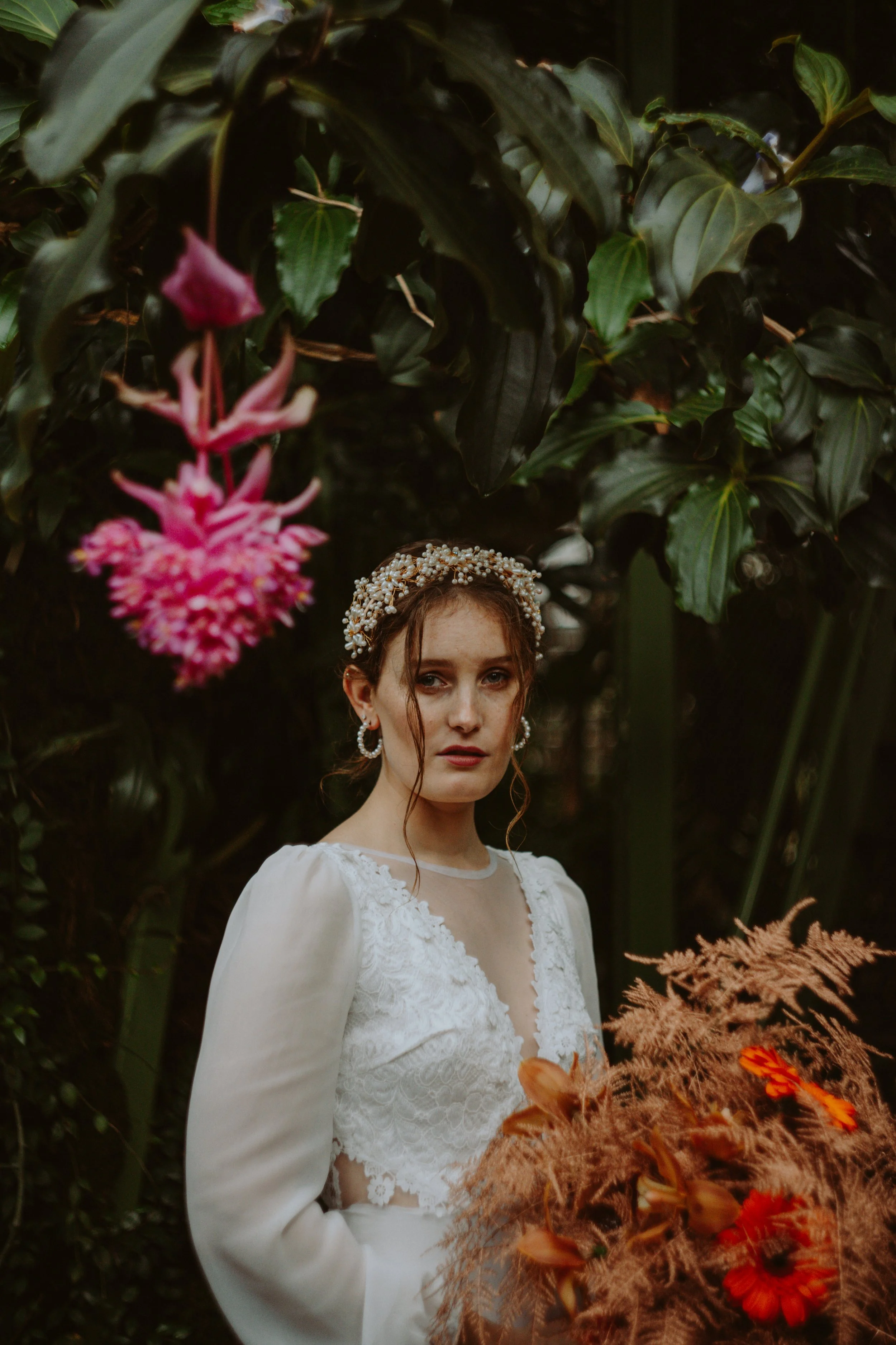 A woman wearing a white dress and a pearl headpiece stands outdoors in front of dark green foliage with pink flowers hanging from a vine above her head.