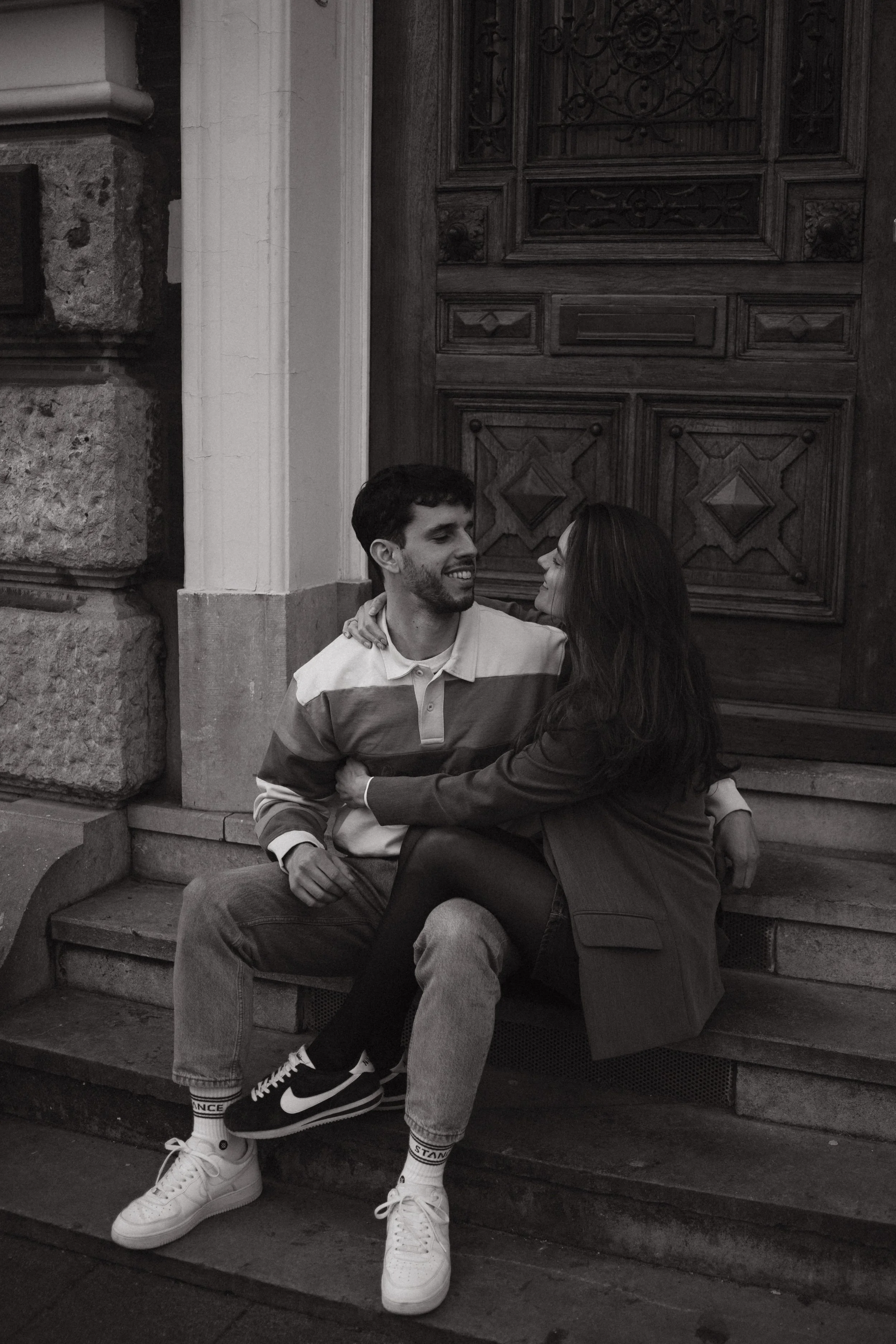 A young man and a young woman are sitting close together on a bench in front of an ornate wooden door. They are smiling and looking into each other's eyes, with the woman's arms around the man's shoulders. The scene is in black and white.