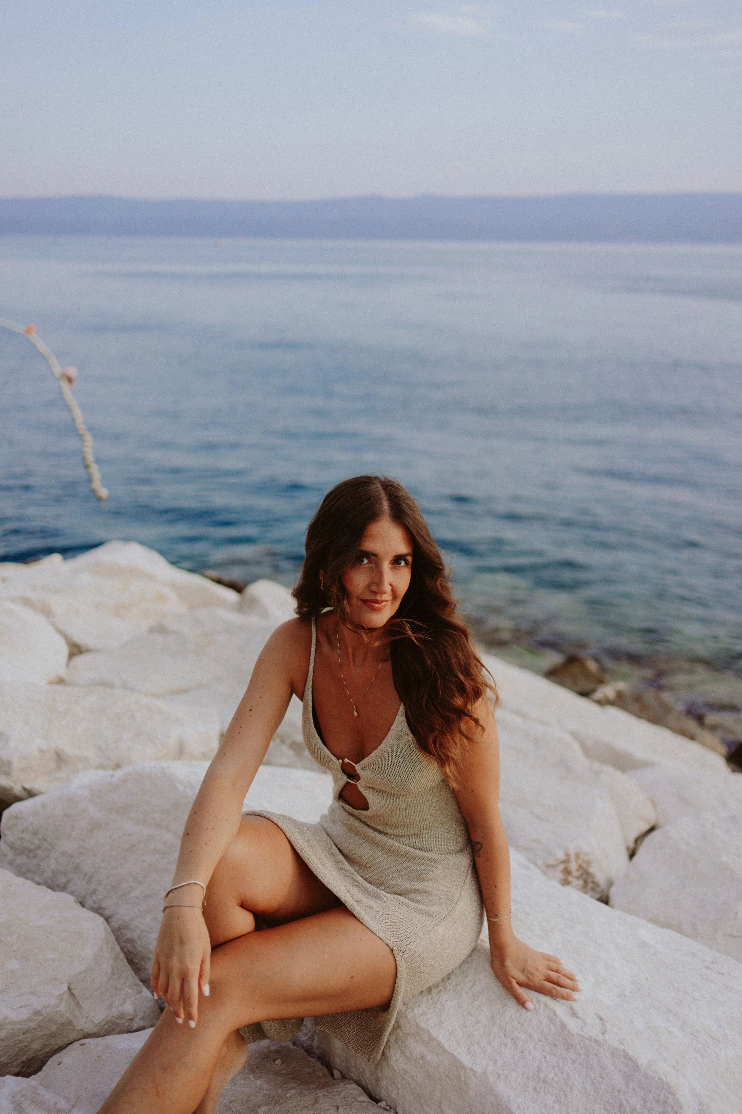Portrait of a  woman with long wavy hair sitting on white rocks near the water at a beach, with a background of calm sea and distant land across the horizon.