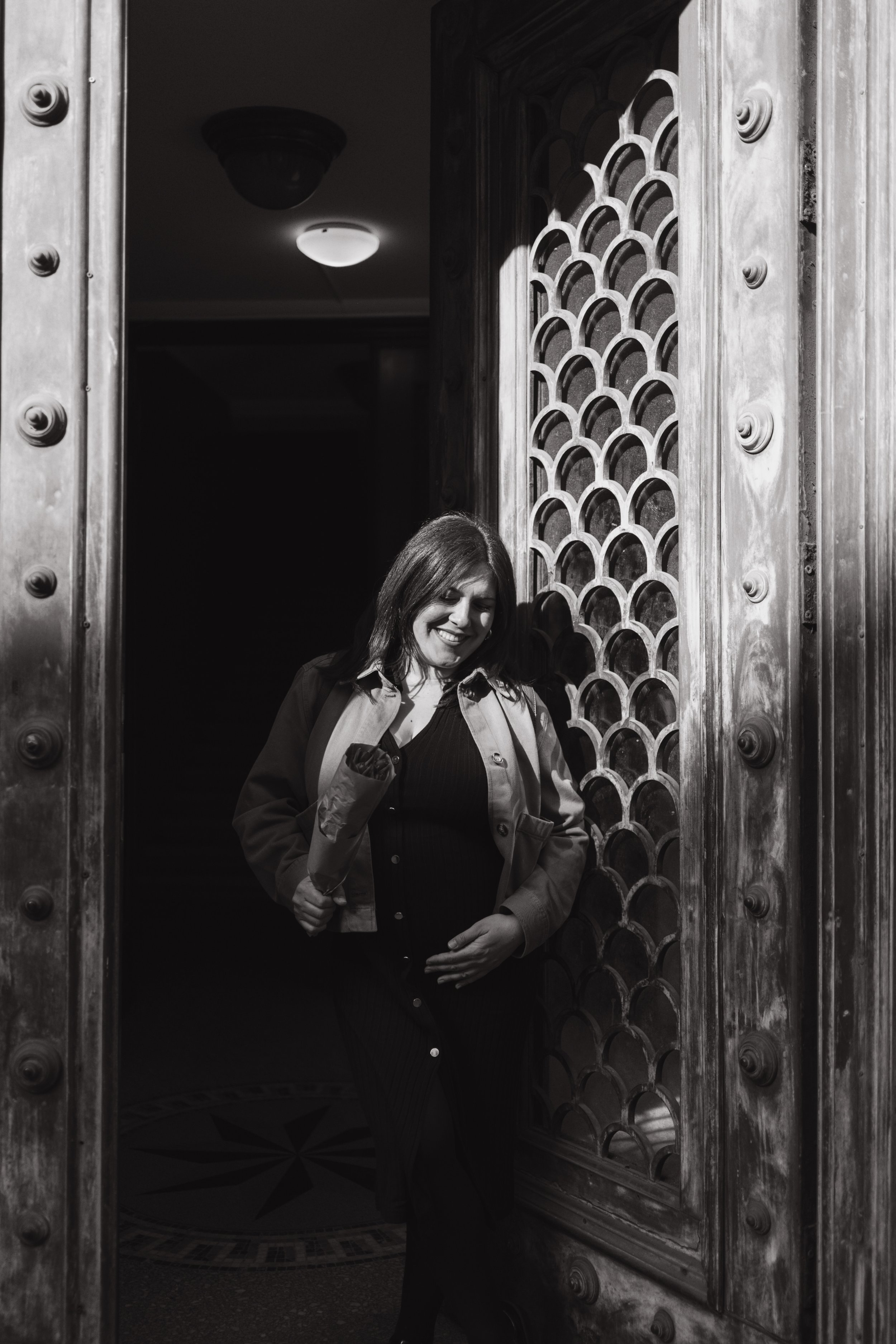 A woman with dark hair and a coat smiling, standing beneath a large open metal door with rivets and a lattice window, inside a dimly lit room or building.