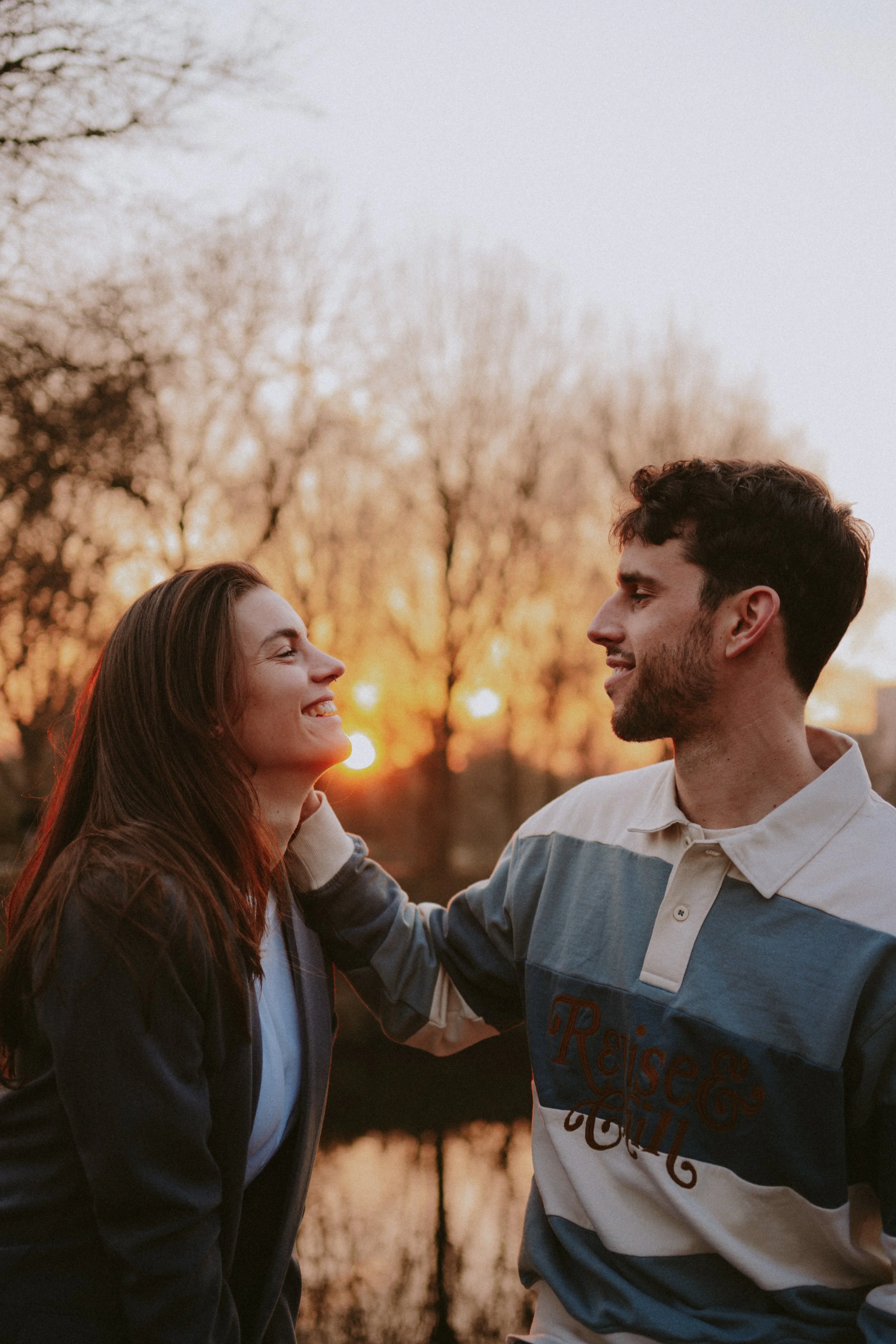 A young woman and man smiling and facing each other outdoors during sunset, with trees and a warm sky in the background.