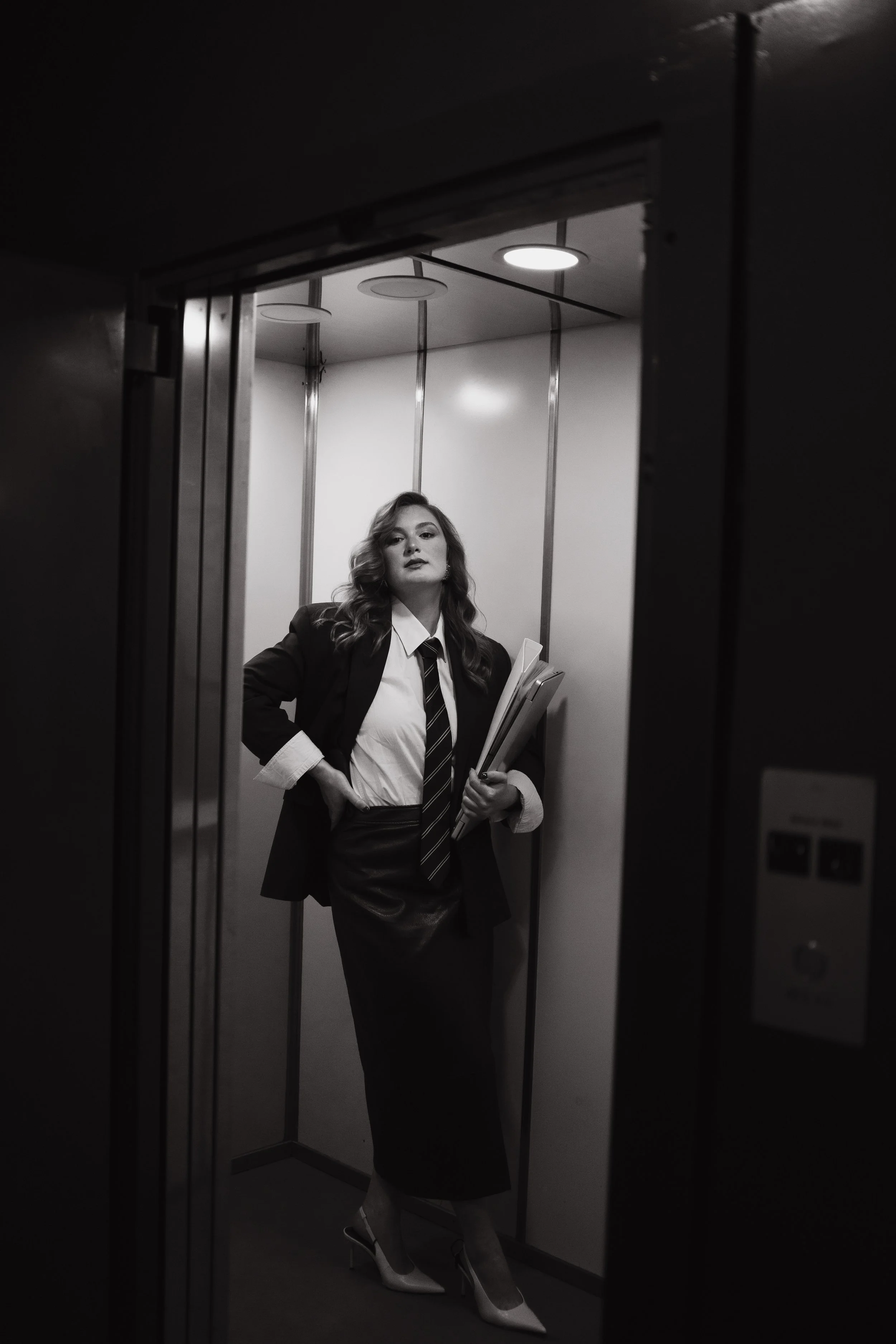 A woman with wavy hair wearing a blazer, white shirt, and tie, standing in an elevator with mirrors on the walls, holding folders, and looking at the camera.