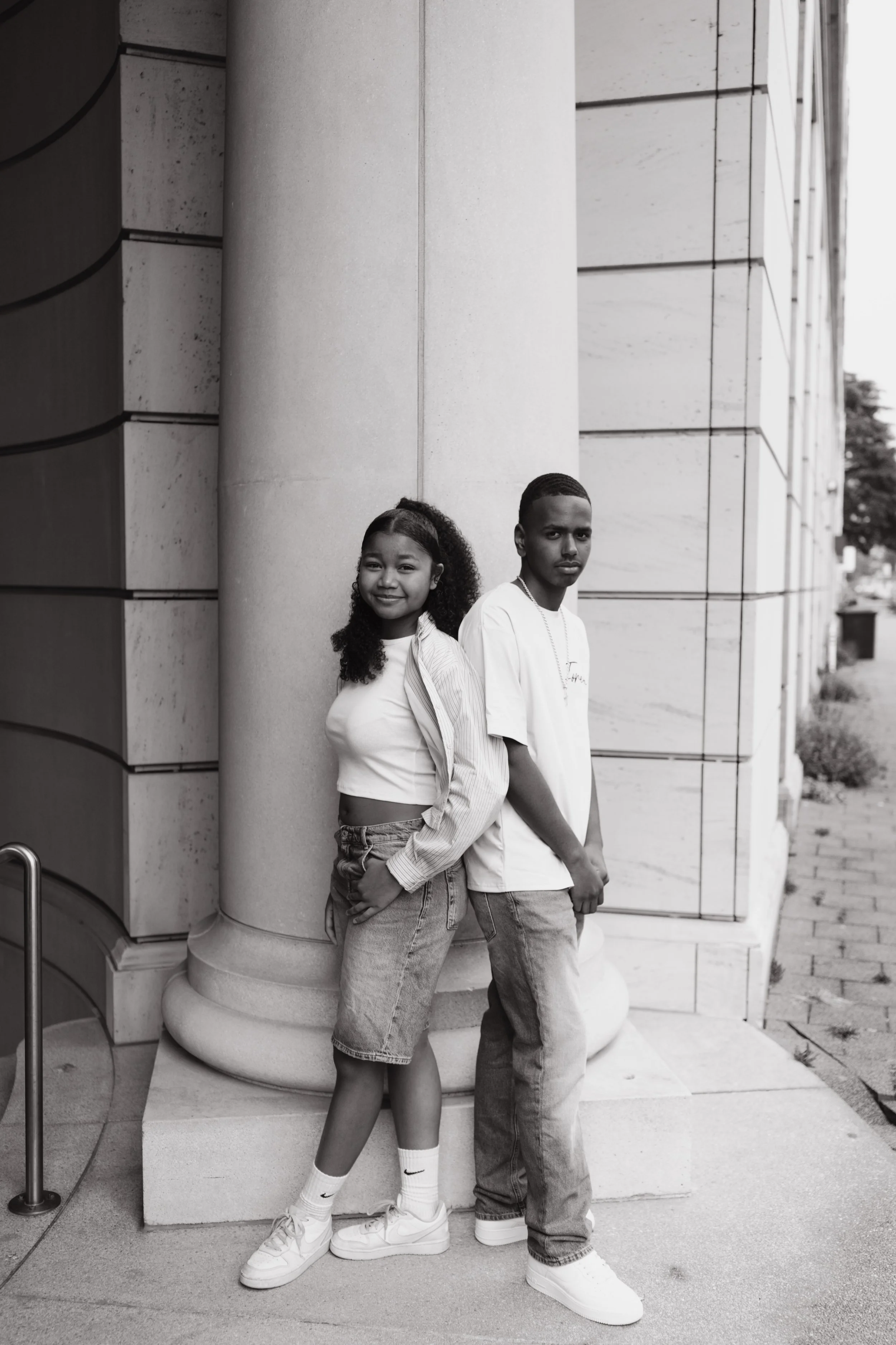 Two young people, a girl and a boy, leaning against a large concrete pillar outside a building. The girl has curly hair, is smiling, and wears a striped shirt, denim shorts, white socks, and sneakers. The boy has short hair, a serious expression, and