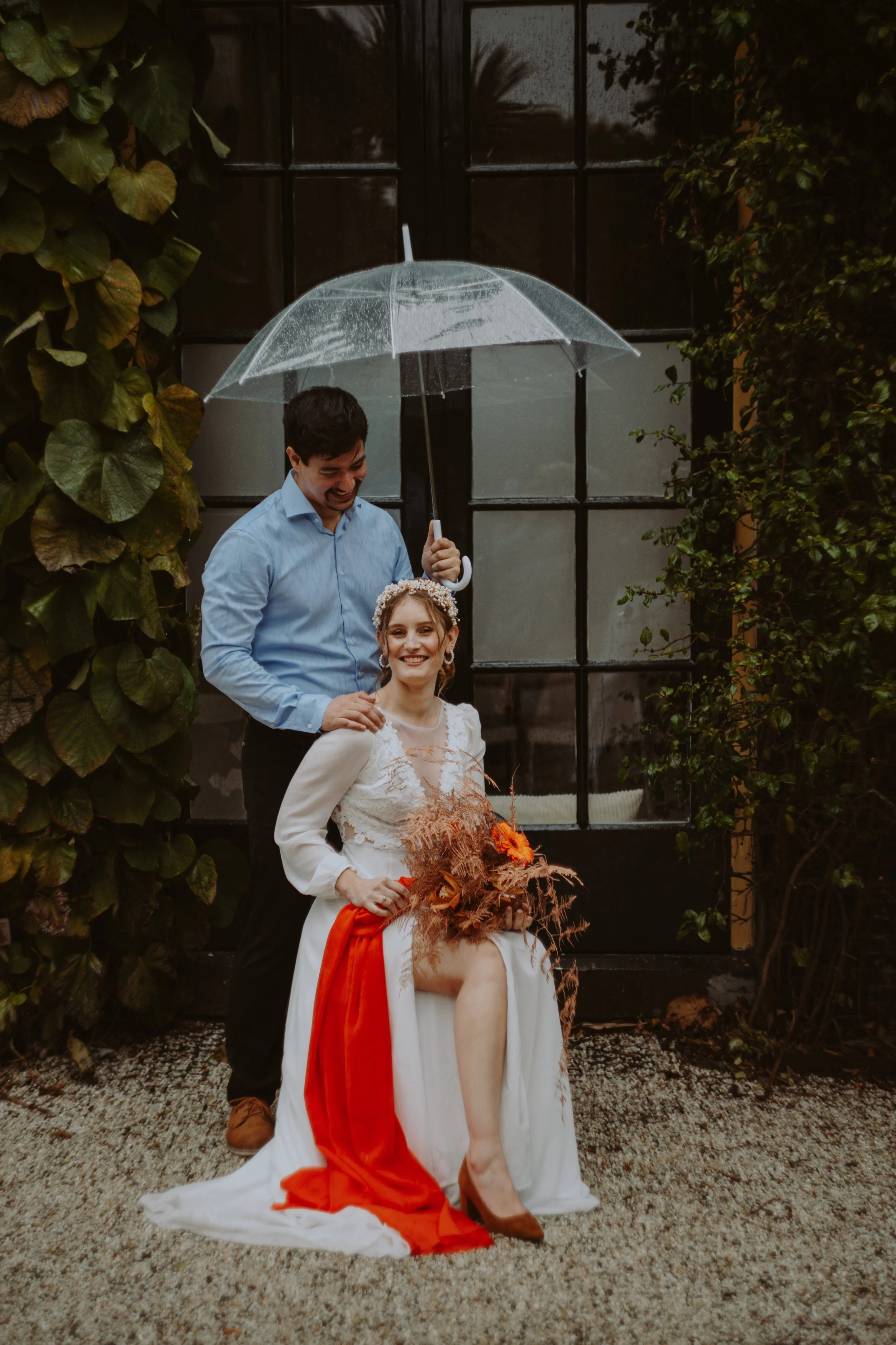 A couple standing outdoors in front of a large black window, with greenery on either side. The woman is sitting on a chair, dressed in a white gown with a red sash, holding a bouquet of flowers, and smiling. The man, wearing a light blue shirt, is st
