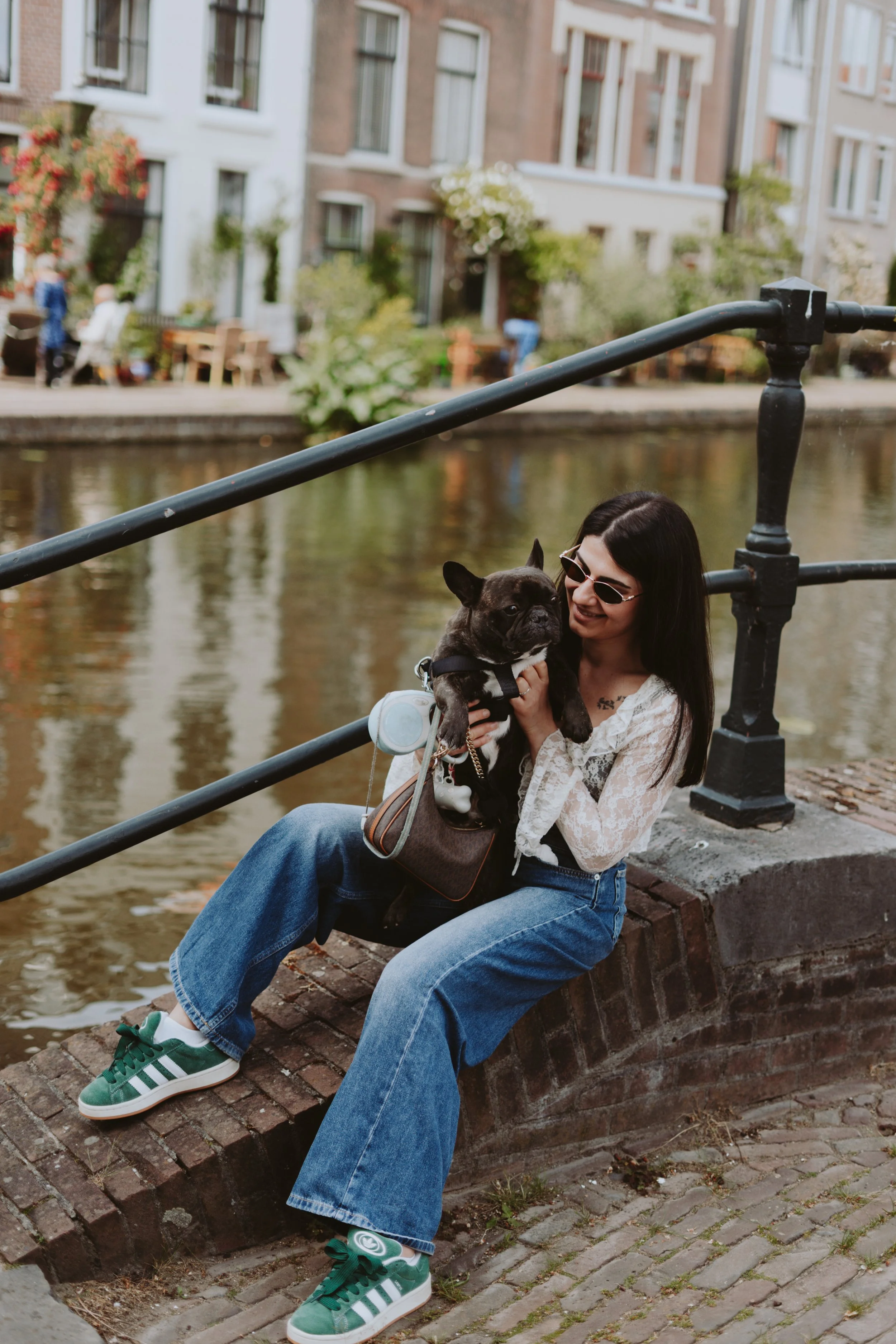 A woman with long, dark hair wearing sunglasses, a white lace top, and blue jeans, sitting on a brick ledge near a canal, holding a small French Bulldog. She has a brown handbag on her lap and is smiling at the dog, which is looking to the side. The 