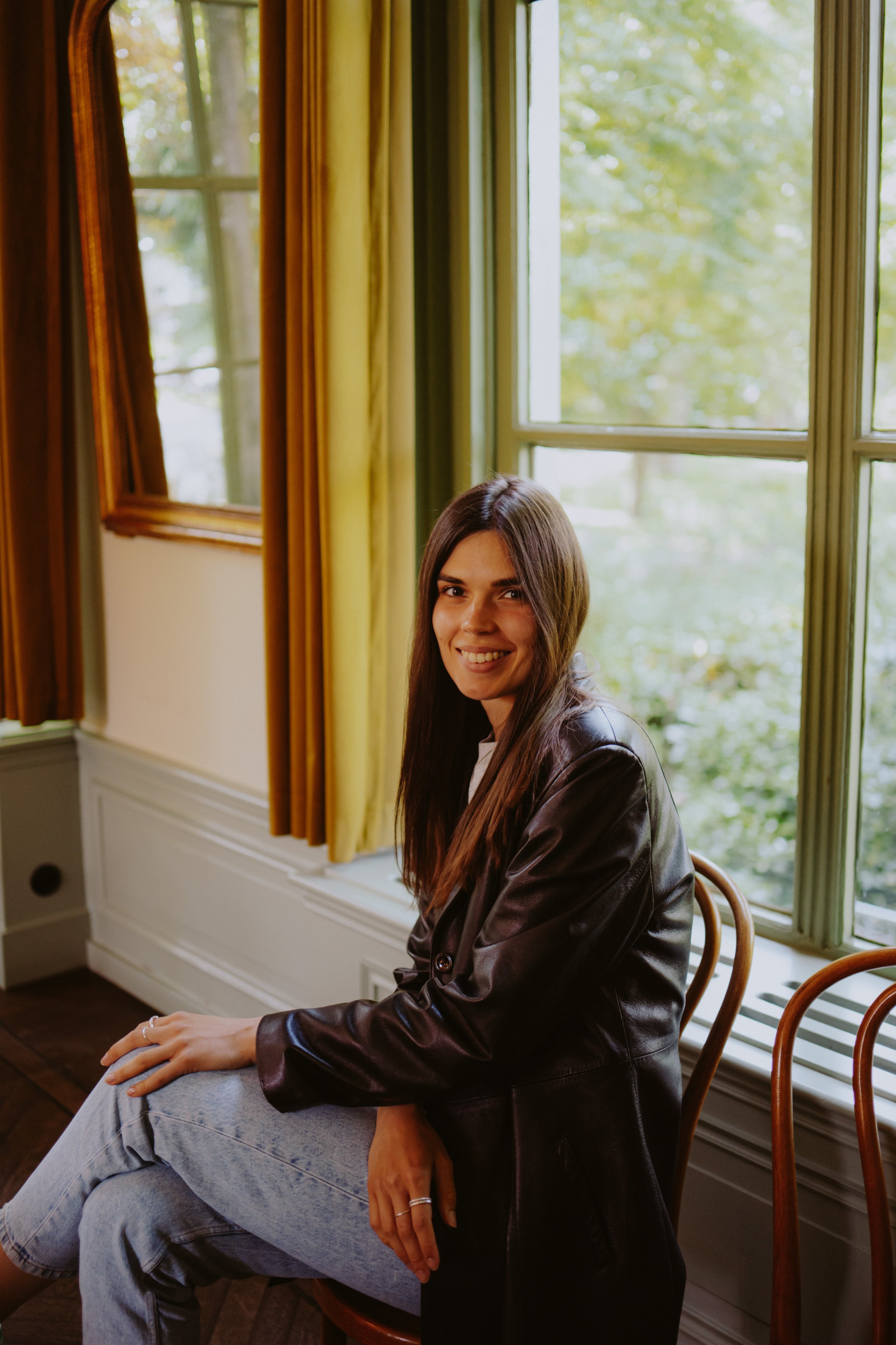 A young woman with long brown hair sitting by a large window with green curtains, smiling and wearing a dark leather jacket and light jeans, with a view of trees outside.