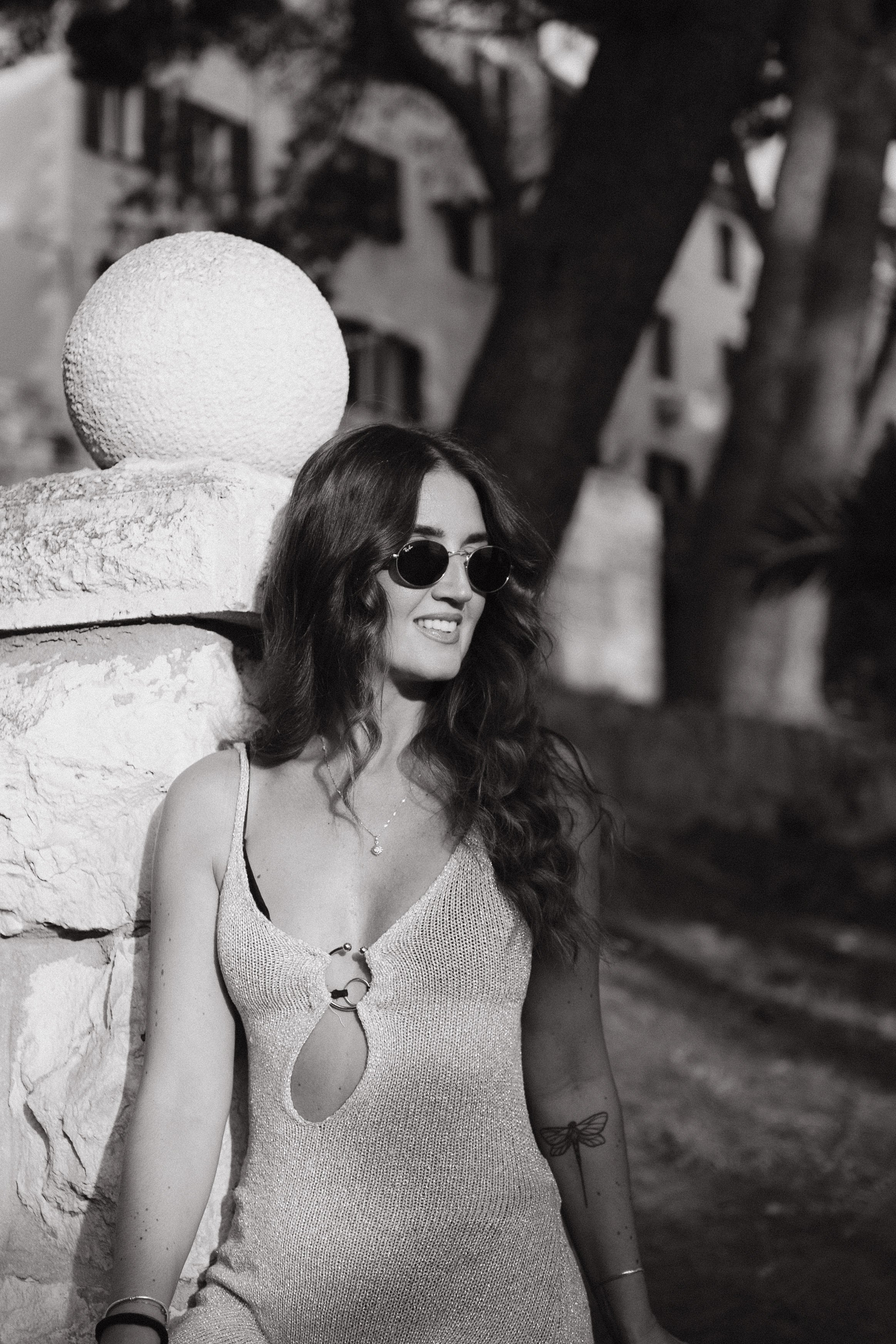A woman with long wavy hair, wearing sunglasses and a sleeveless dress with cutouts, standing next to a textured stone wall with a large spherical sculpture, trees, and buildings in the background.