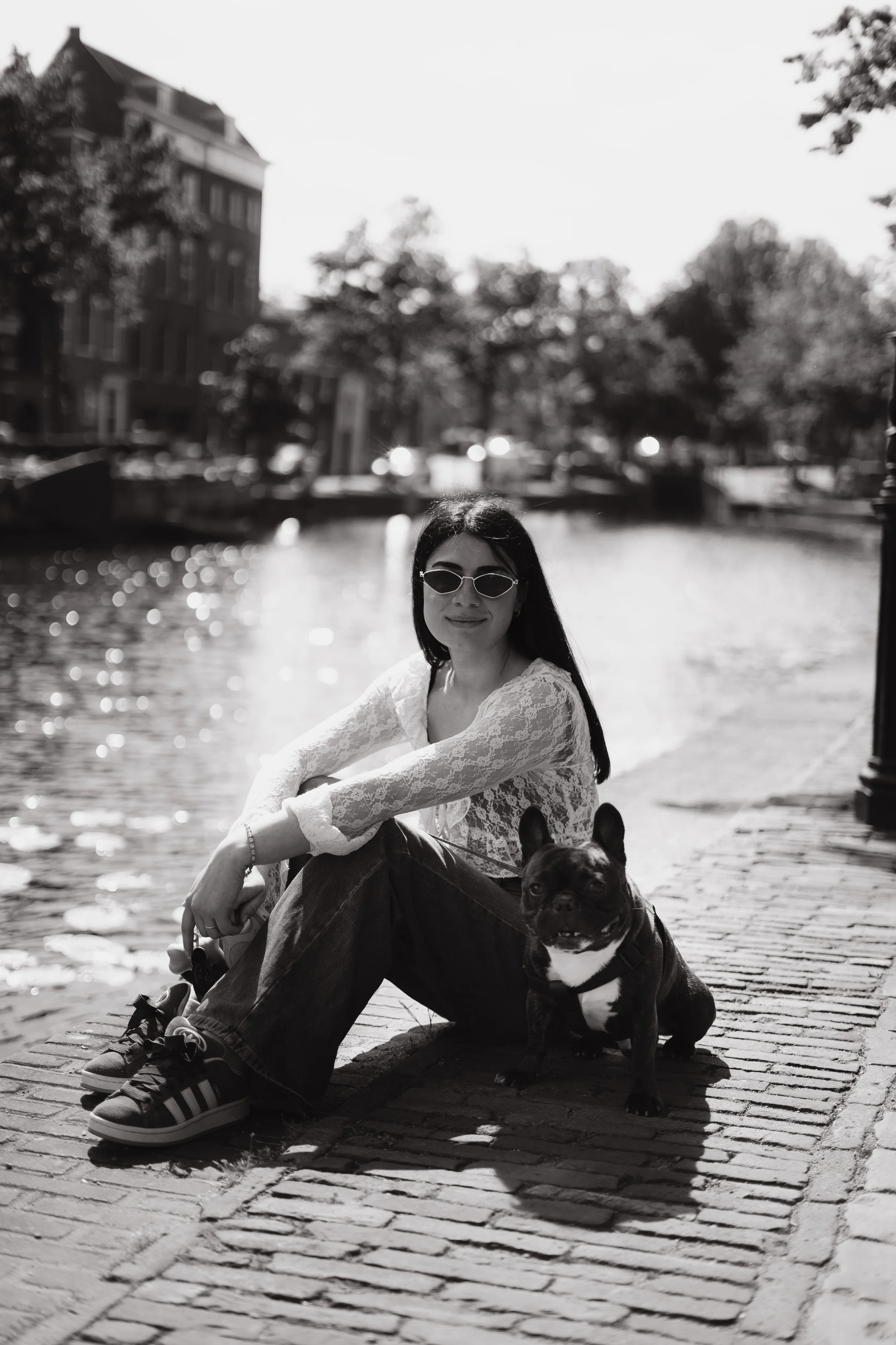 A woman wearing sunglasses and a lace top sitting on a brick sidewalk by a body of water, with a French bulldog sitting beside her. There are trees and buildings in the background.