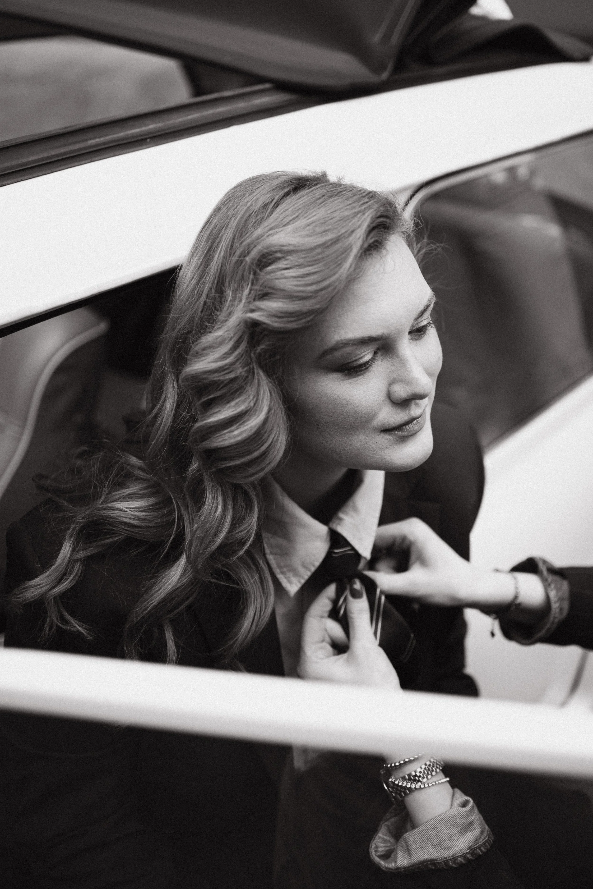 Black and white photo of a woman with wavy hair, wearing a collared shirt and blazer, sitting in a car with the window down, looking to her side.