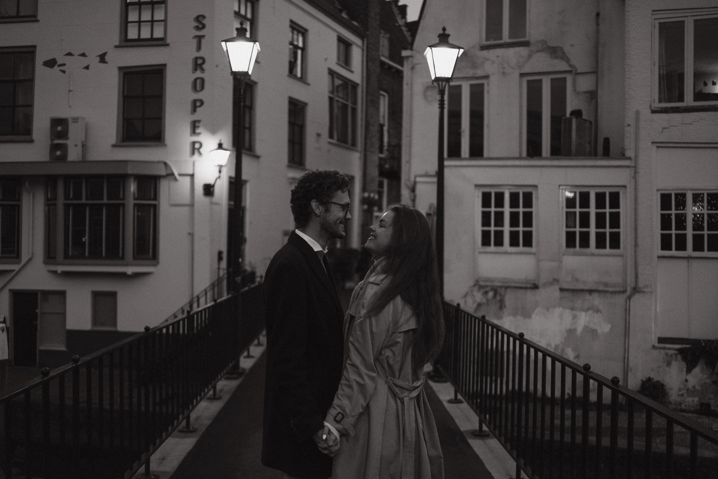 A black and white photo of a couple holding hands and smiling at each other on a bridge at night, with city buildings and street lamps in the background.