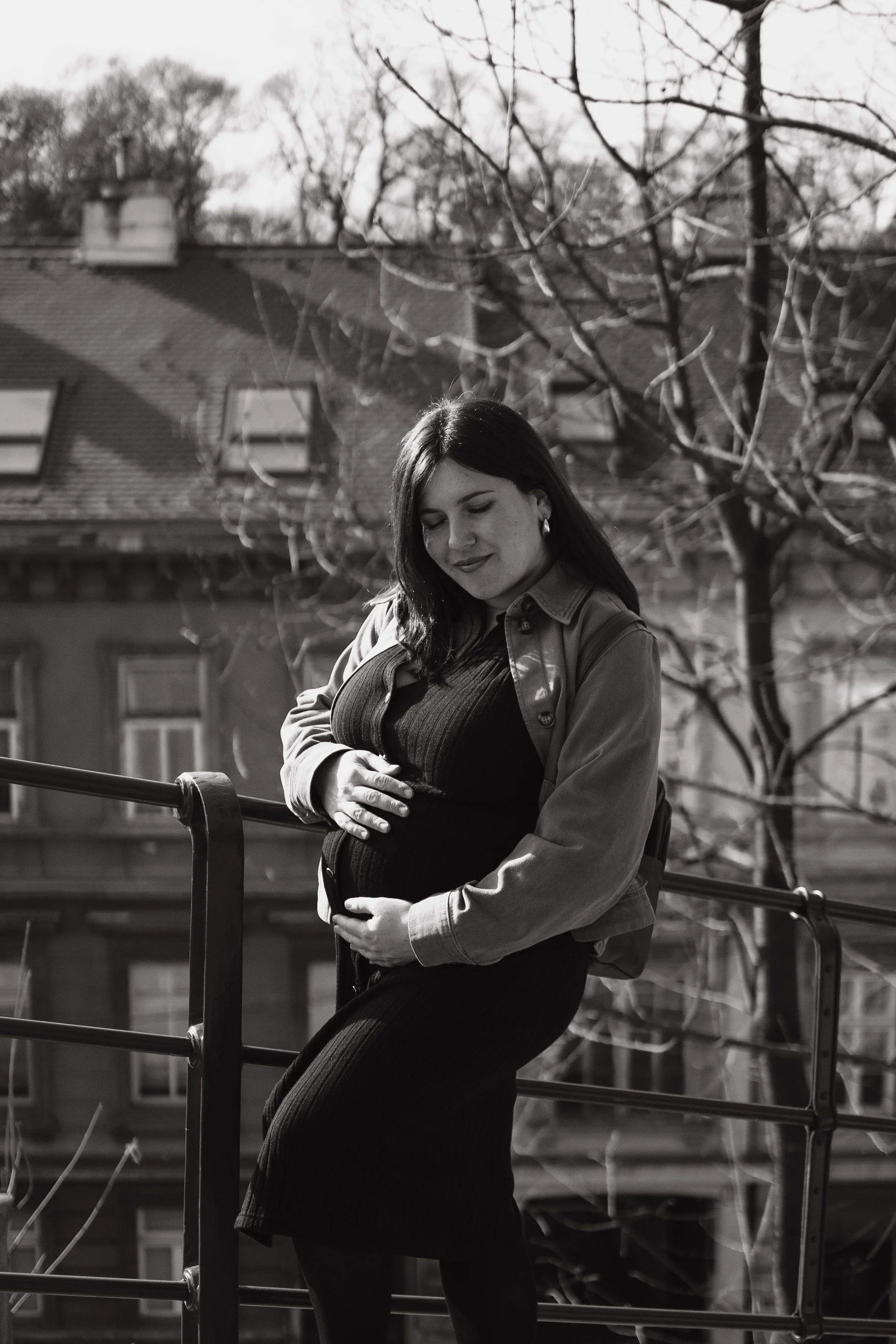Black and white photo of a woman with dark hair, wearing earrings, standing outside in front of buildings and leafless trees, smiling gently, with her arms crossed over her stomach.