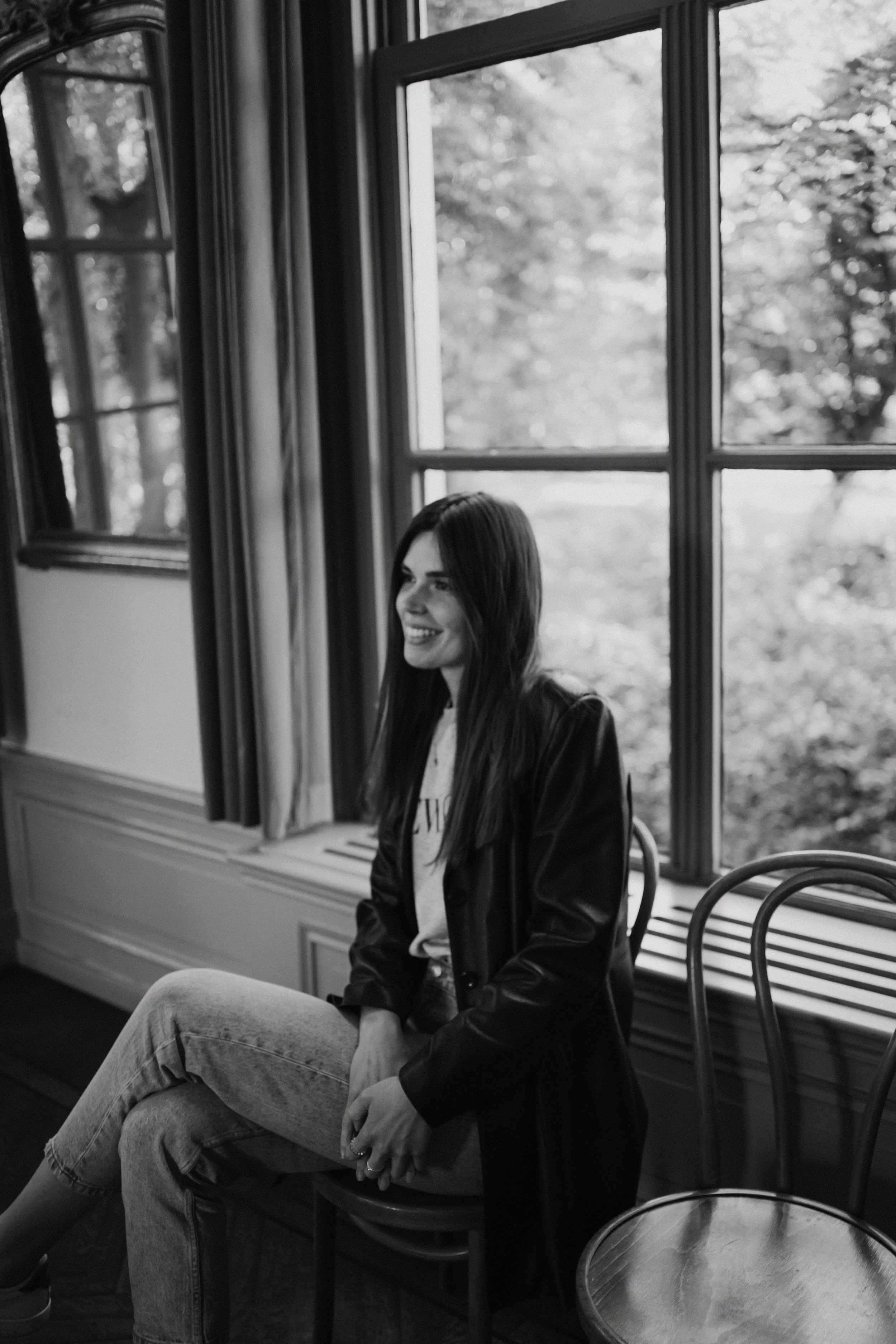 A woman with long dark hair sitting on a chair in front of large window with curtains, smiling and looking out, in a room with wooden floors and round table.
