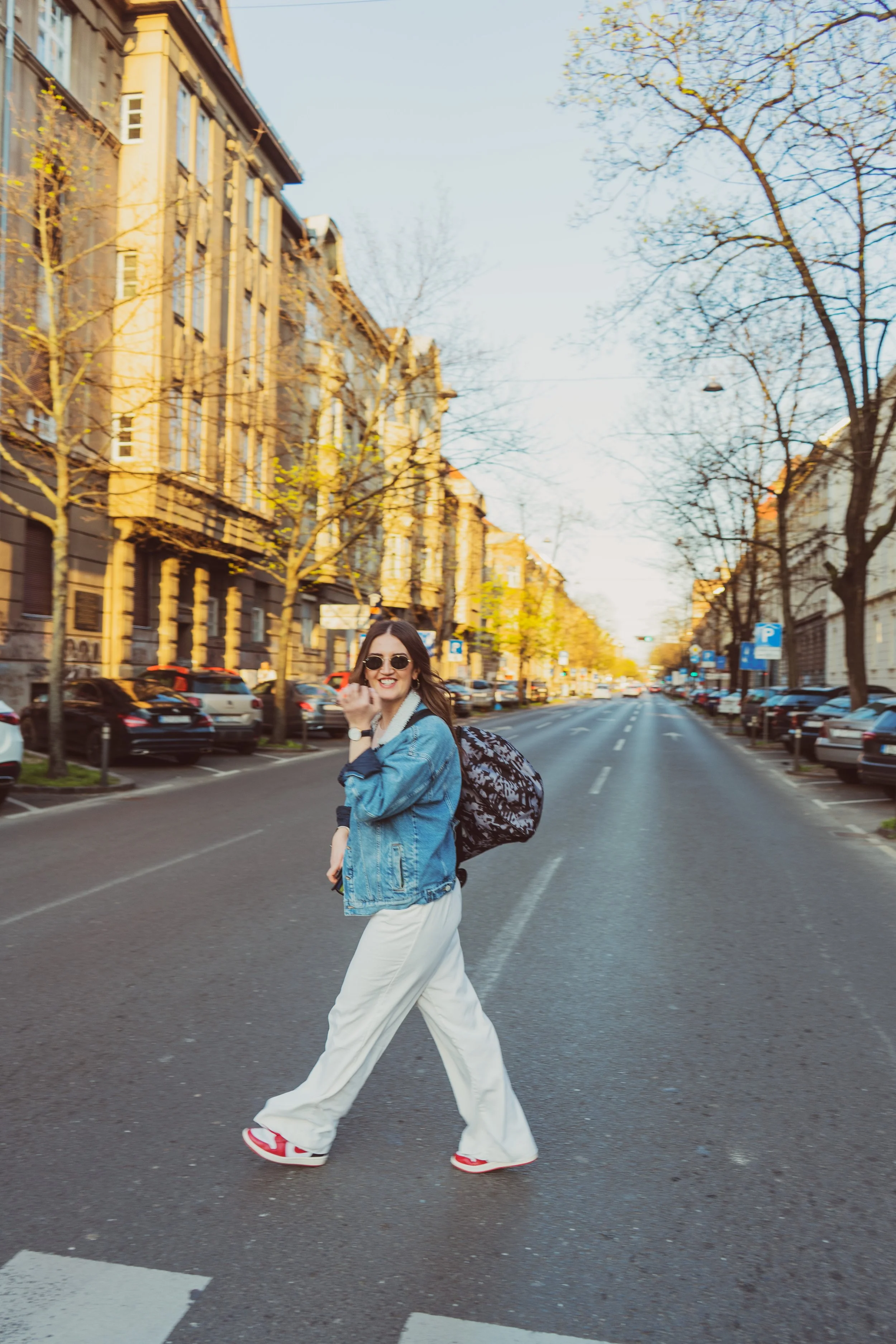 A young woman with sunglasses and a denim jacket crossing a city street at a pedestrian crosswalk, smiling and holding her hand up in a playful gesture, with cars parked along the street and buildings on either side.