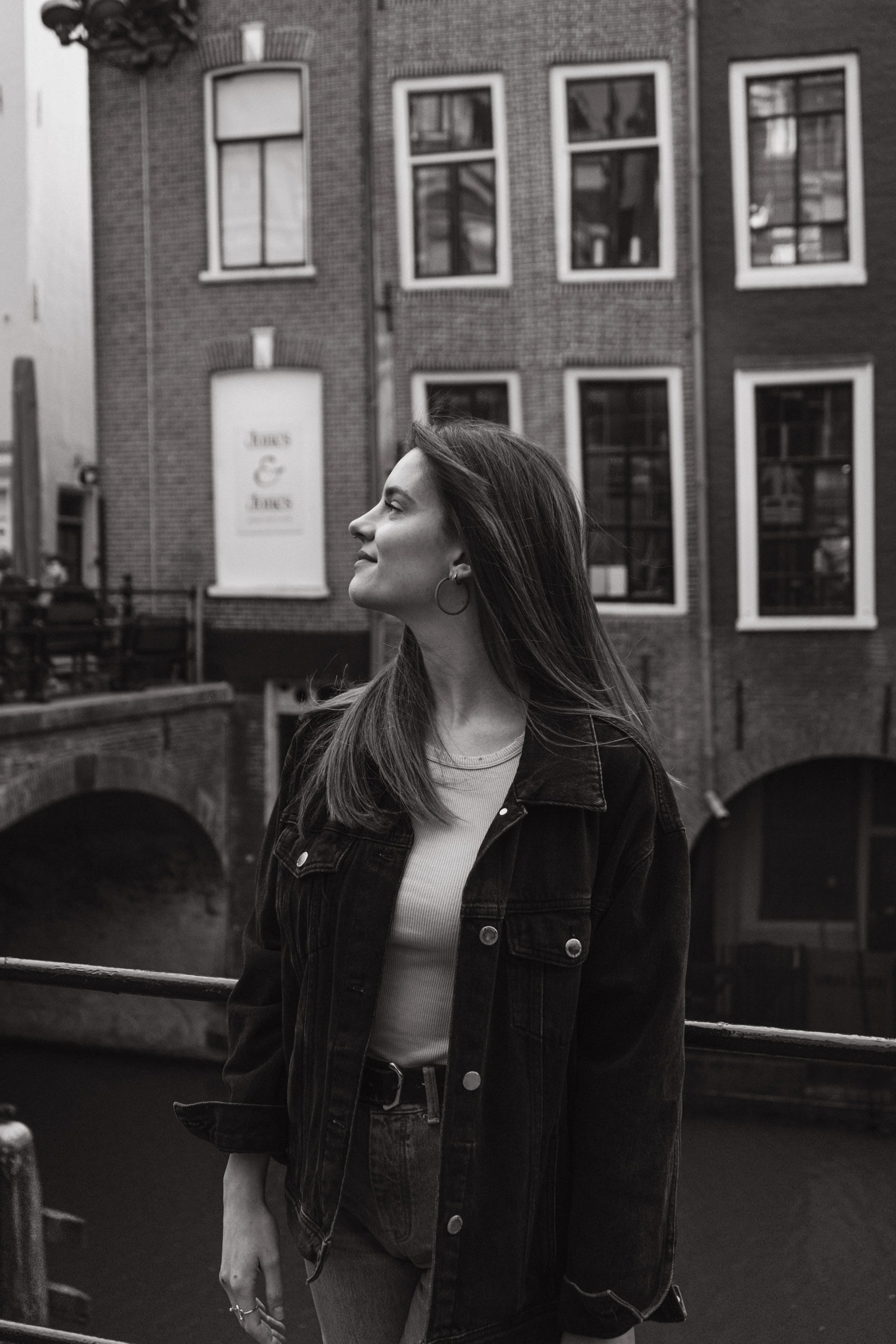 A woman with long hair wearing hoop earrings, a white top, and a denim jacket standing outdoors near a brick building with multiple windows and a sign that reads 'love & life' in the background.