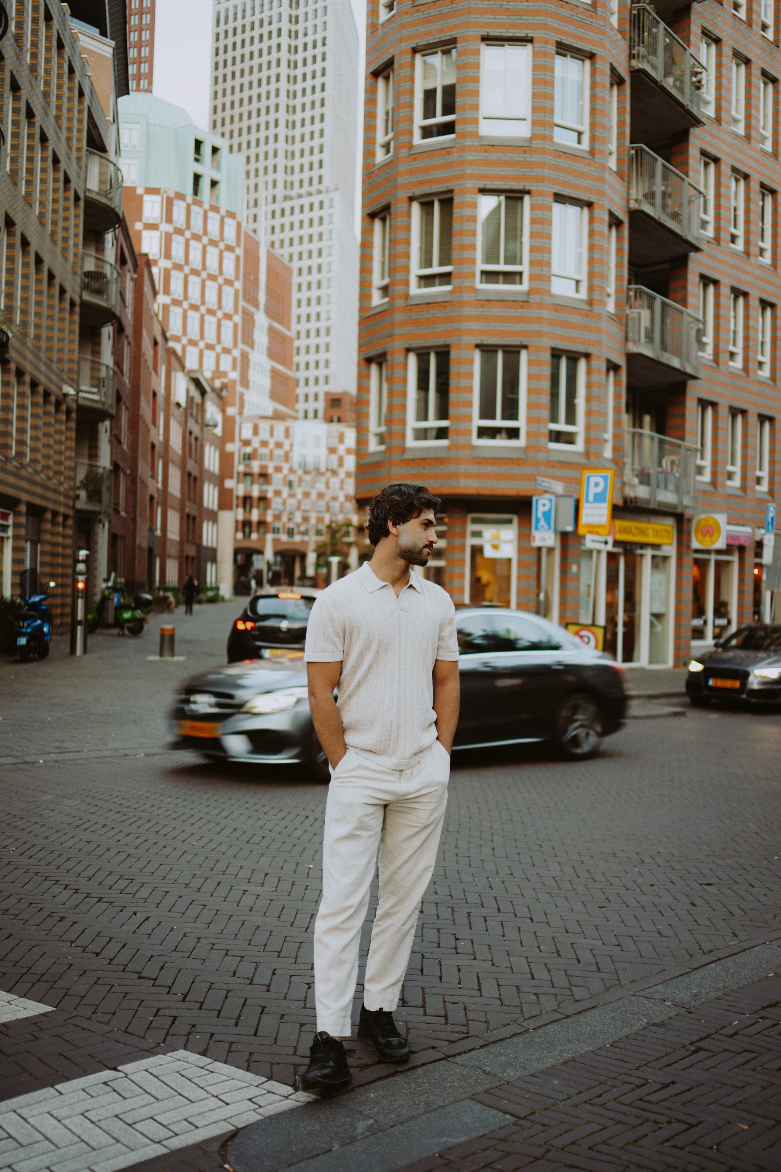 A man with dark curly hair, a beard, wearing a white polo shirt and white pants, standing on a city street with tall modern buildings in the background. Vehicles are moving past him.
