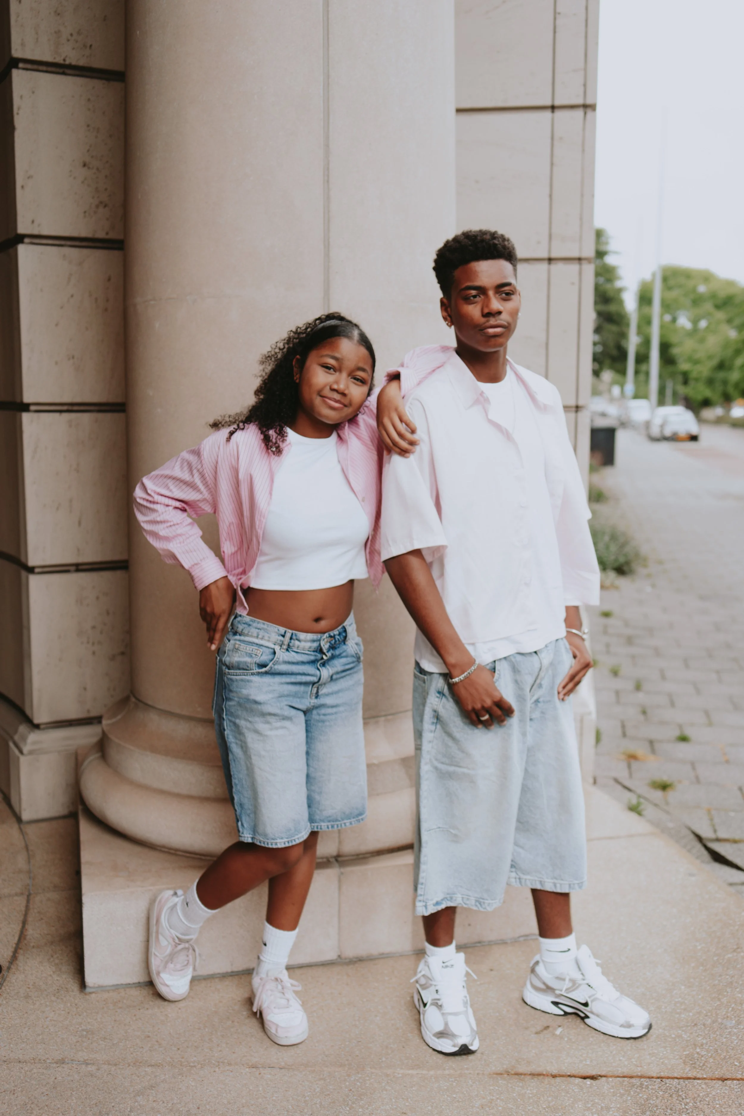 Two young people, a girl and a boy, standing against a building exterior. The girl is wearing a pink striped shirt over a white crop top, denim shorts, and white sneakers. The boy is dressed in a light pink shirt, baggy denim shorts, and white sneake