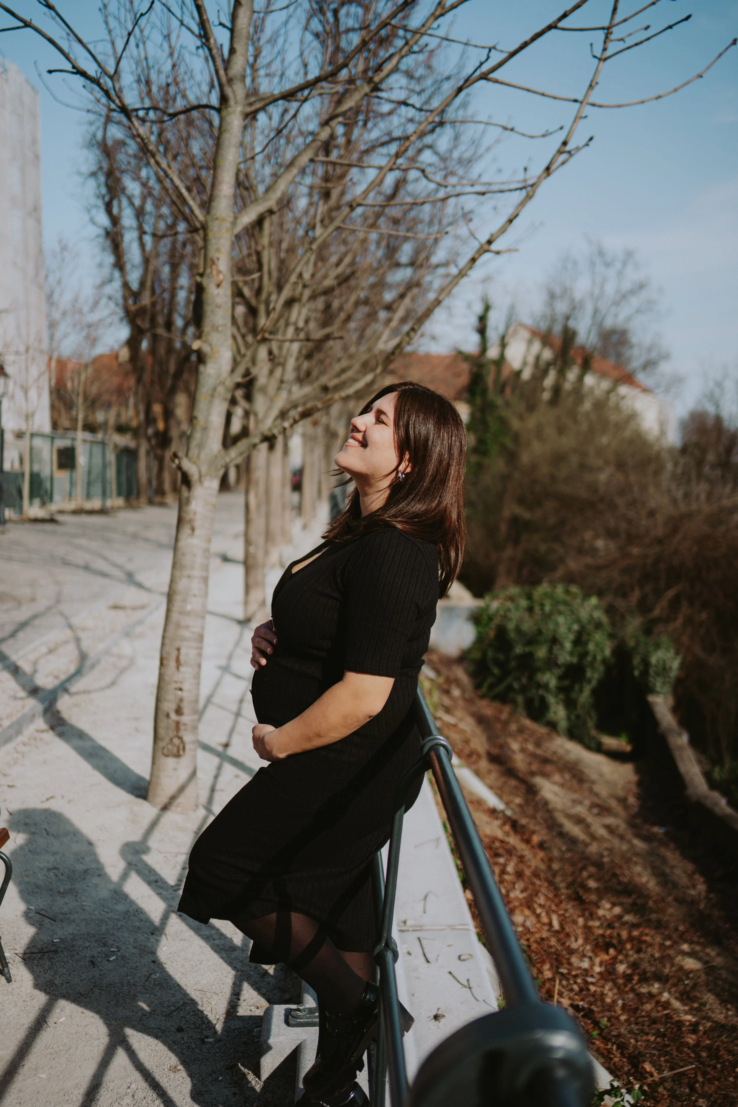 Woman in black dress leaning on railing outdoors in park, smiling with eyes closed, trees in background.