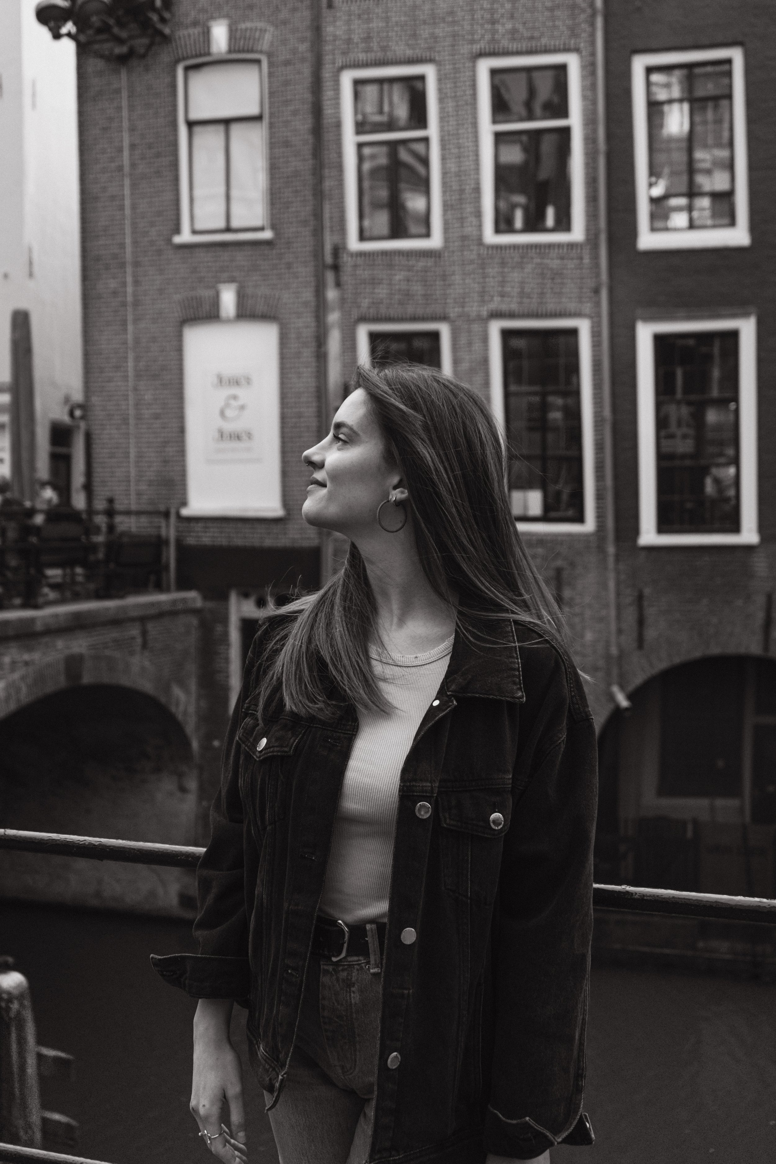 A woman with long hair wearing earrings, a light-colored top, and a denim jacket standing near a river with buildings in the background.