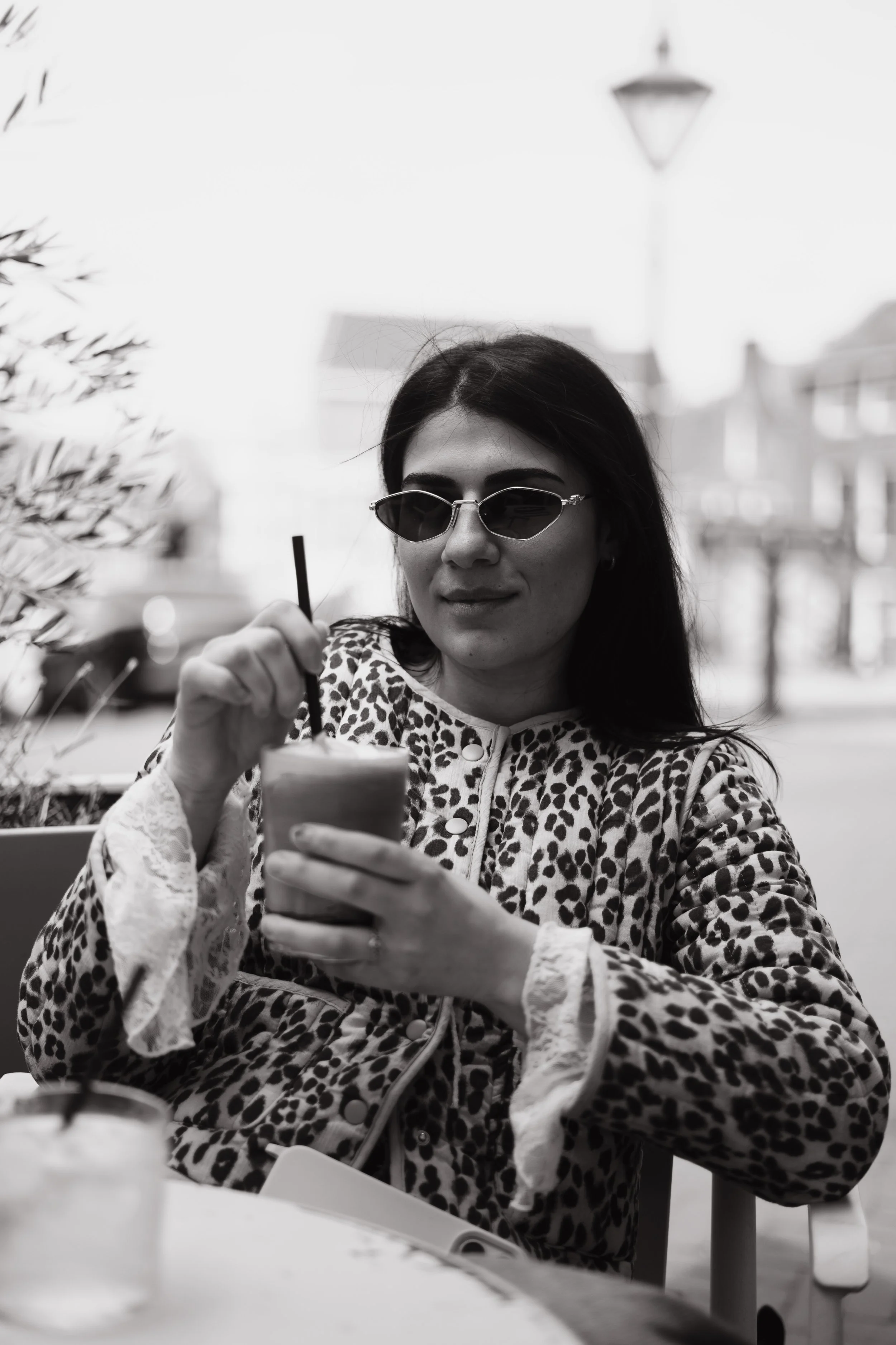 A woman with dark hair wearing sunglasses and a leopard print coat is sitting at a table. She is holding a spoon and taking a bite of food from a cup or glass.