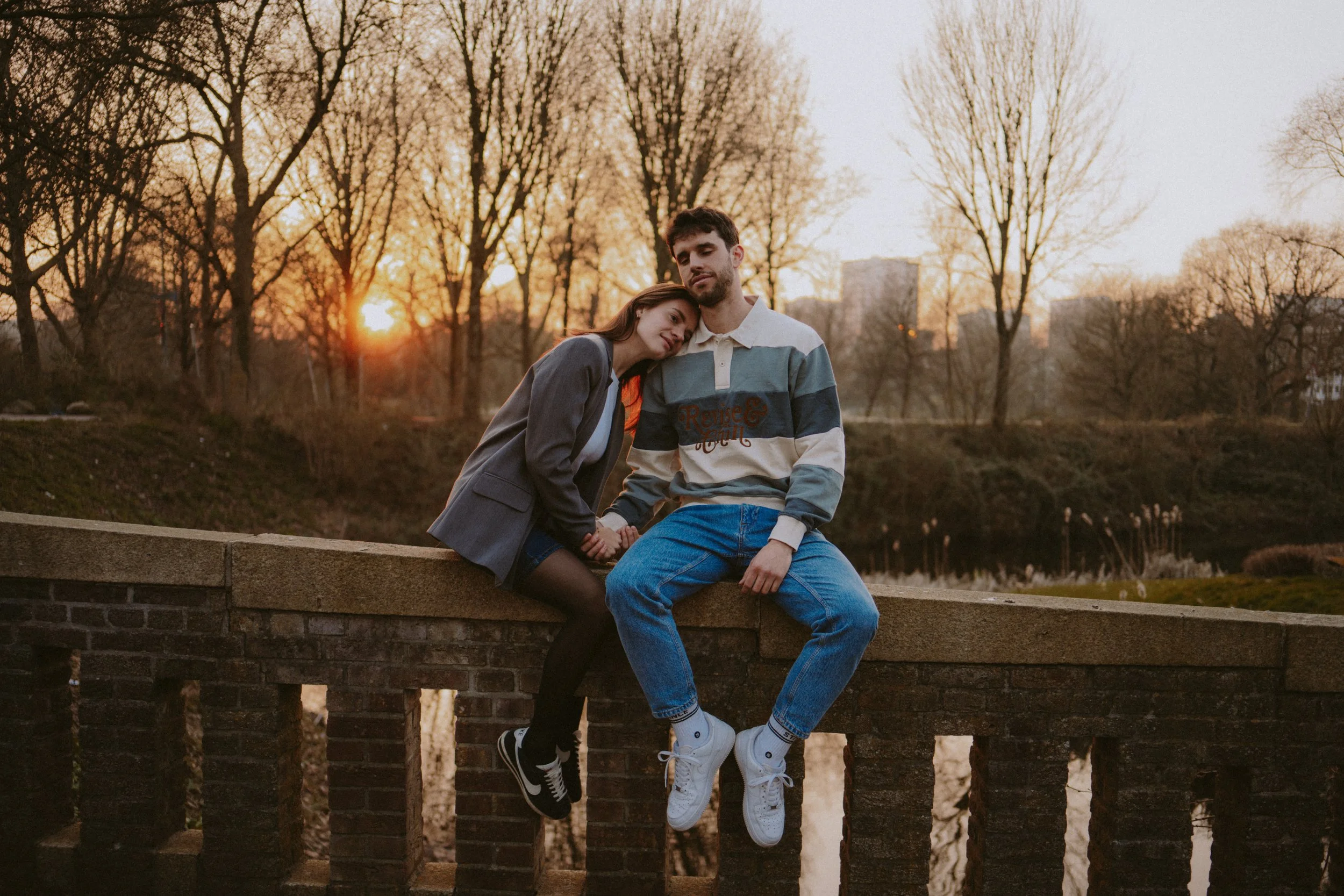 A young couple sitting on a brick wall by a river during sunset, with trees in the background.