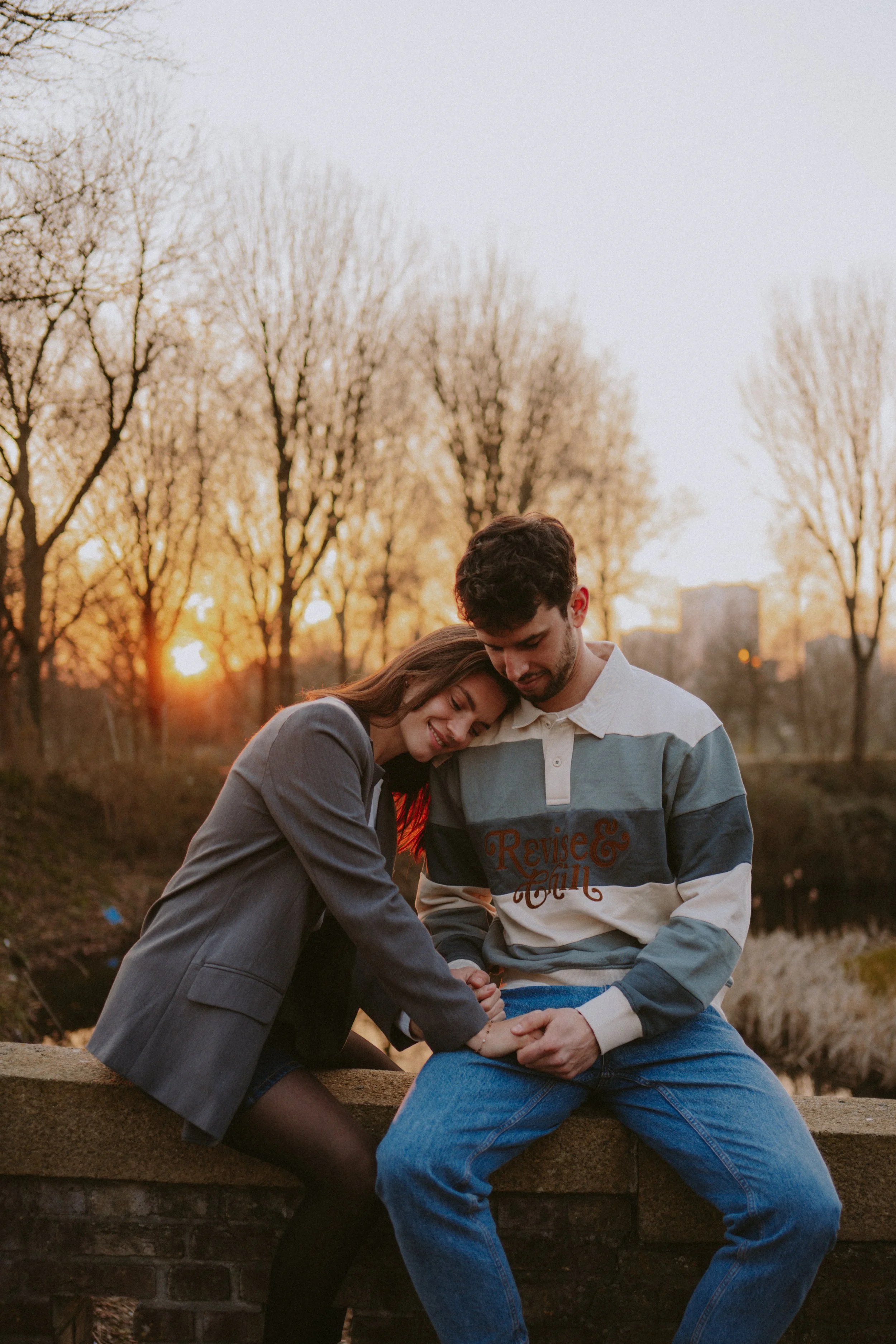A couple sitting close together on a stone ledge by a lake at sunset, with trees in the background, sharing a tender moment.