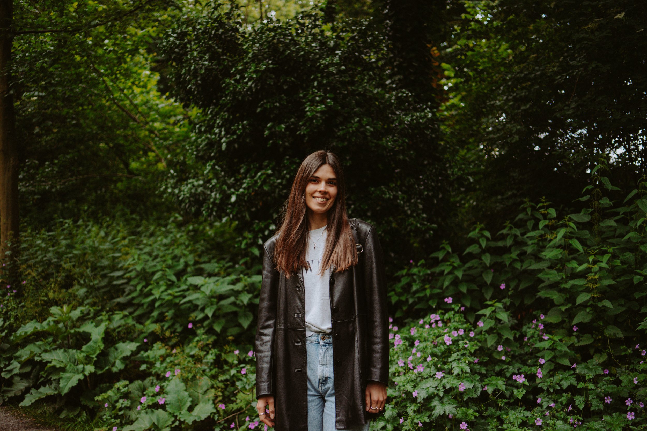 A young woman with long brown hair, wearing a black leather jacket and a white T-shirt, smiling outdoors in front of dense green foliage and purple flowers.