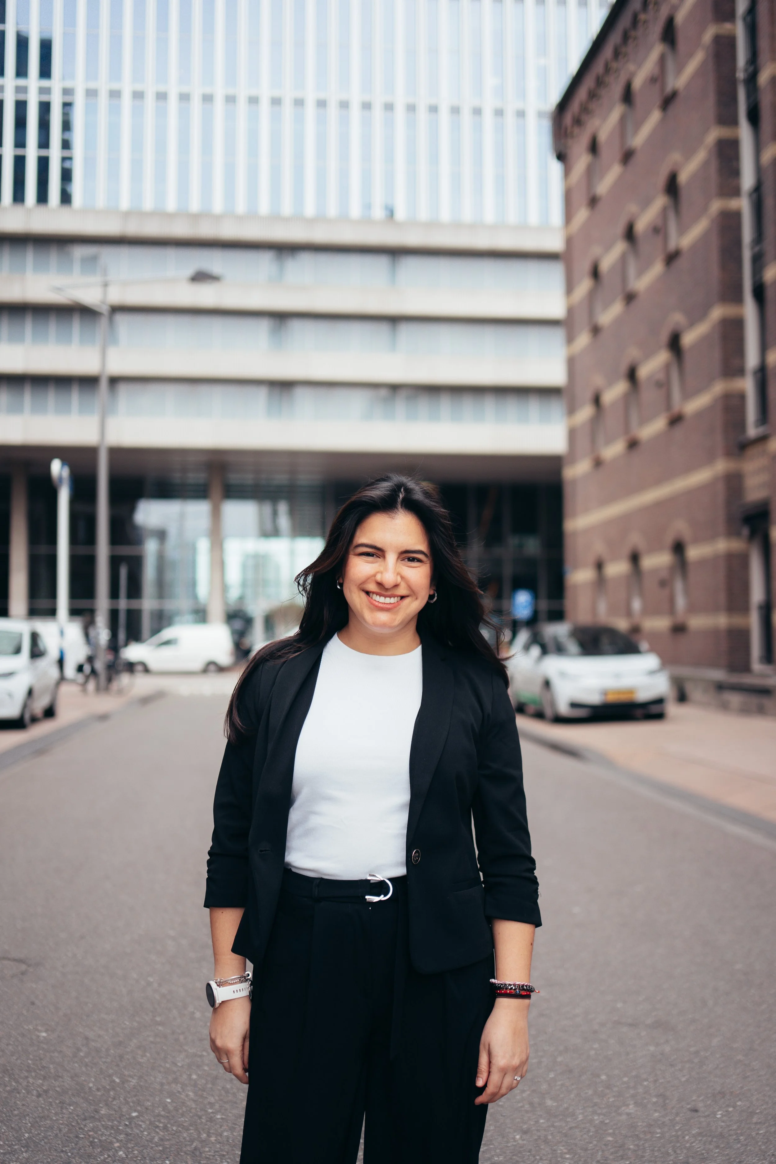 A woman with dark hair smiling and wearing a black blazer and pants, white shirt, standing on a city street with modern glass and brick buildings and parked cars in the background.