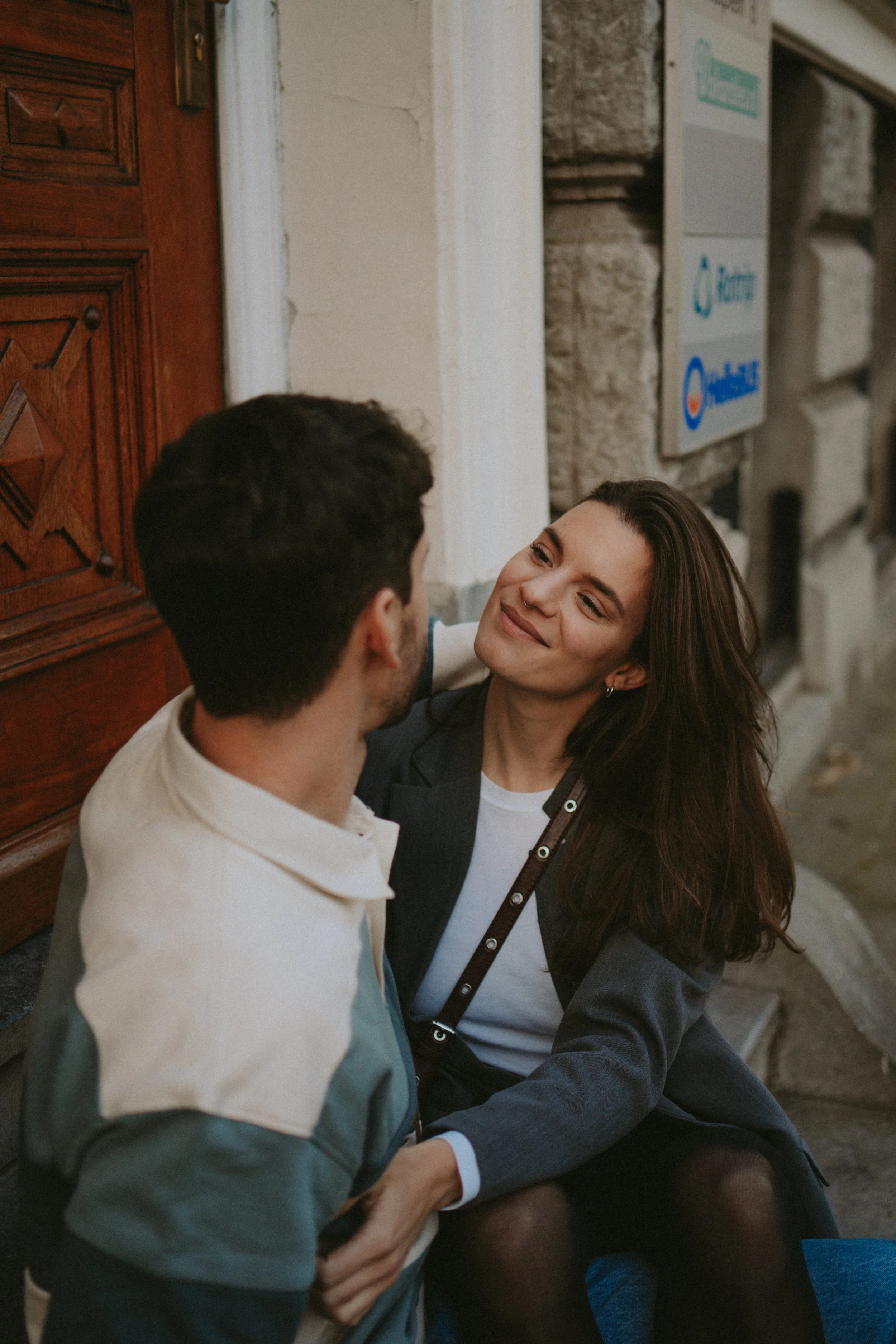 A couple sitting on the ground, holding hands, facing each other and smiling, in an urban setting next to a stone and brick building.