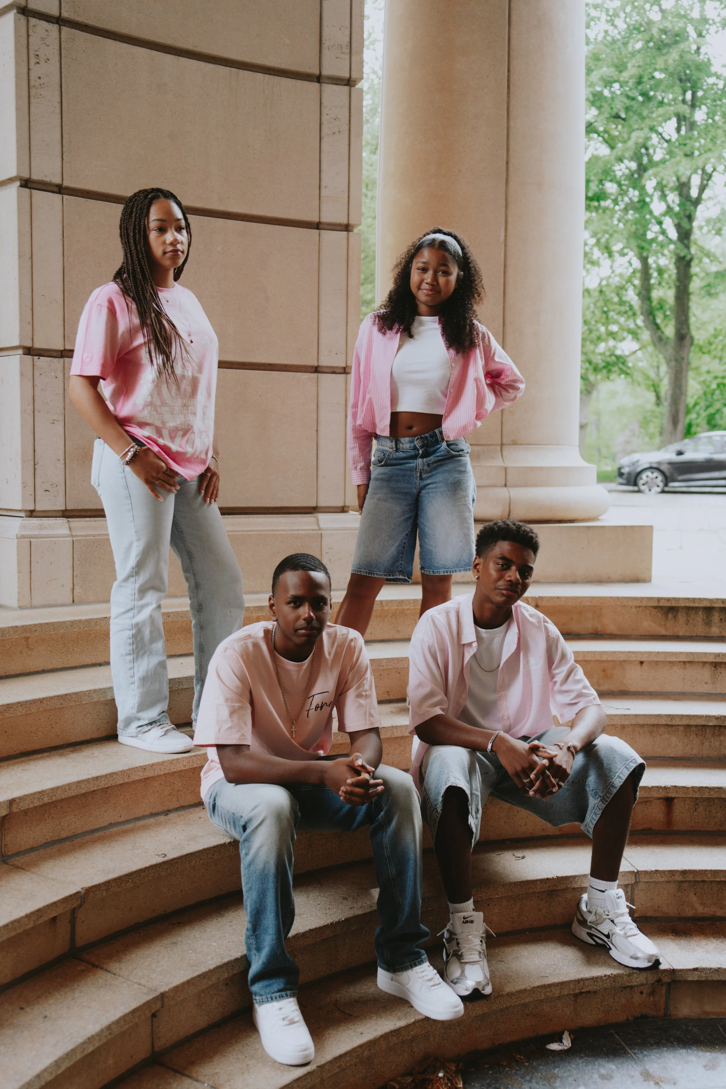 A group of six young people posing on the steps outside a large building with columns, with trees and a car in the background.