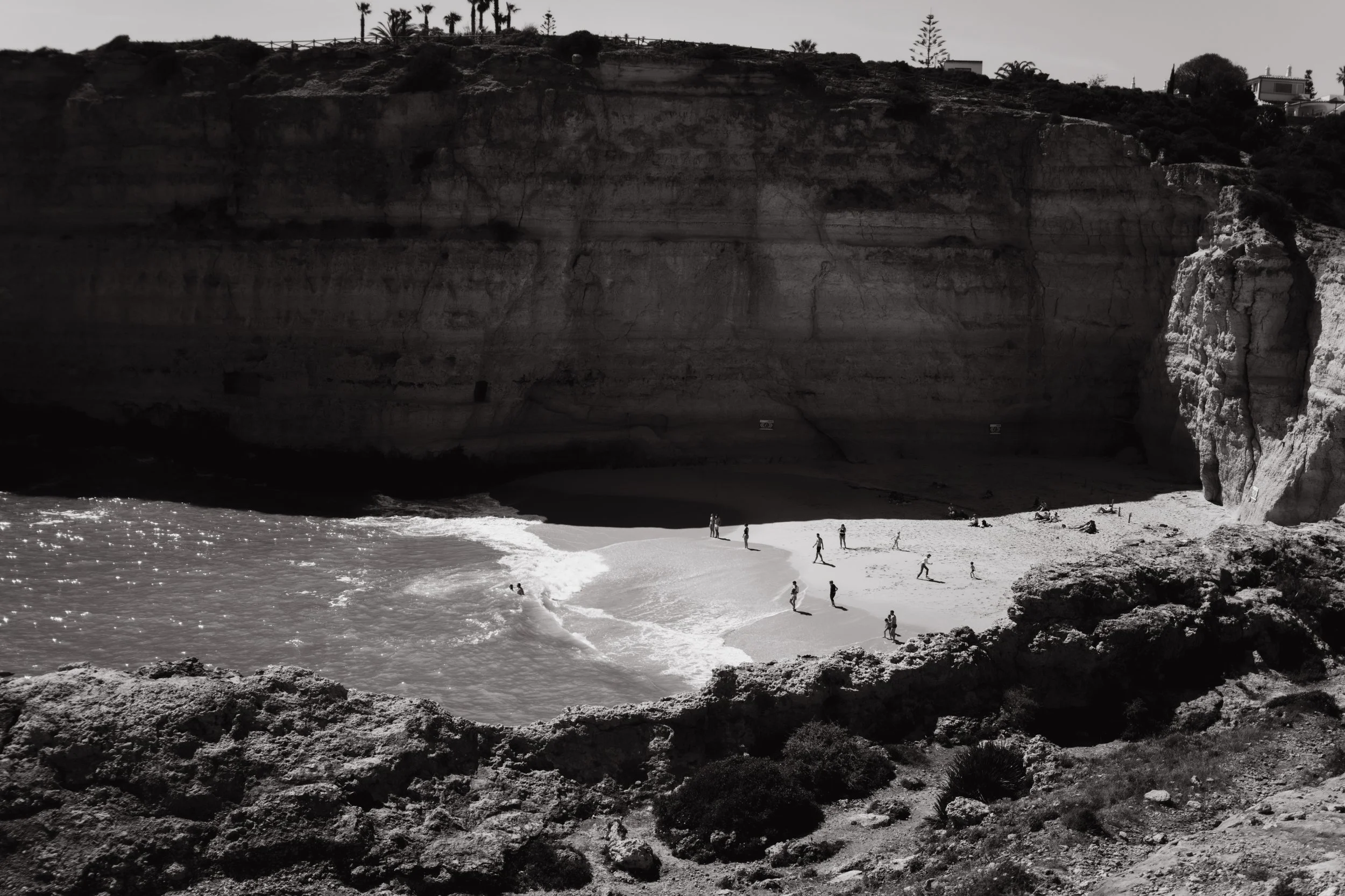 Travel photography of people walking on a sandy beach near a high cliff with trees and houses on top, ocean waves at the shore.