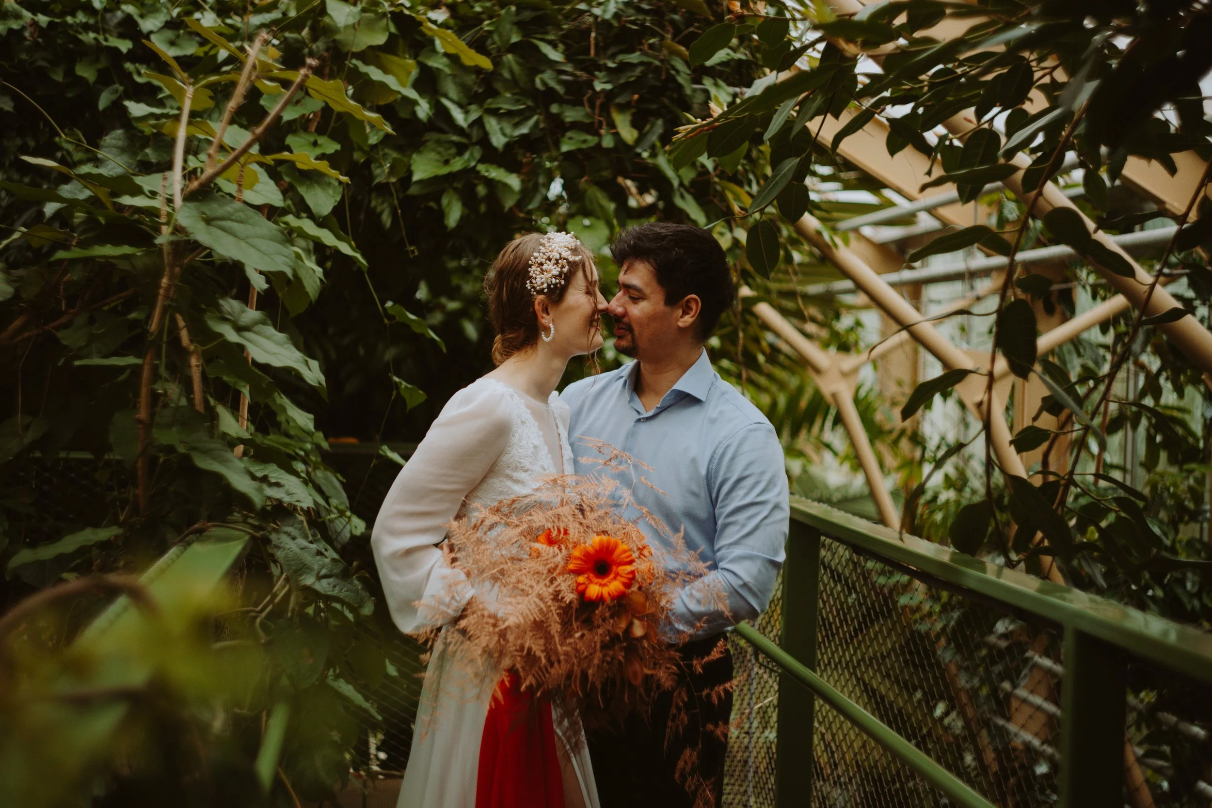 A couple is standing close together, nose to nose, inside a greenhouse surrounded by lush green plants. The woman is wearing a white dress with lace details and a floral headpiece, holding a bouquet of orange flowers. The man is in a light blue shirt. They are sharing an intimate moment, smiling gently at each other.