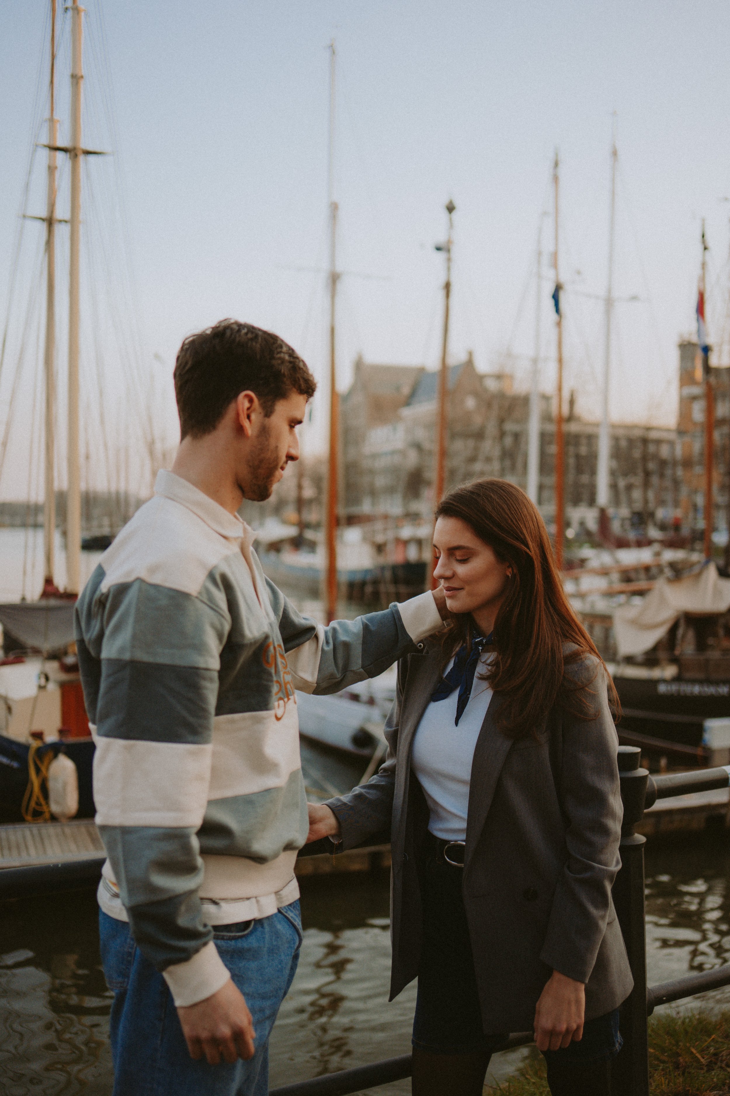 A man and woman standing near a waterfront with boats and sailboats in the background, engaging in a tender moment.