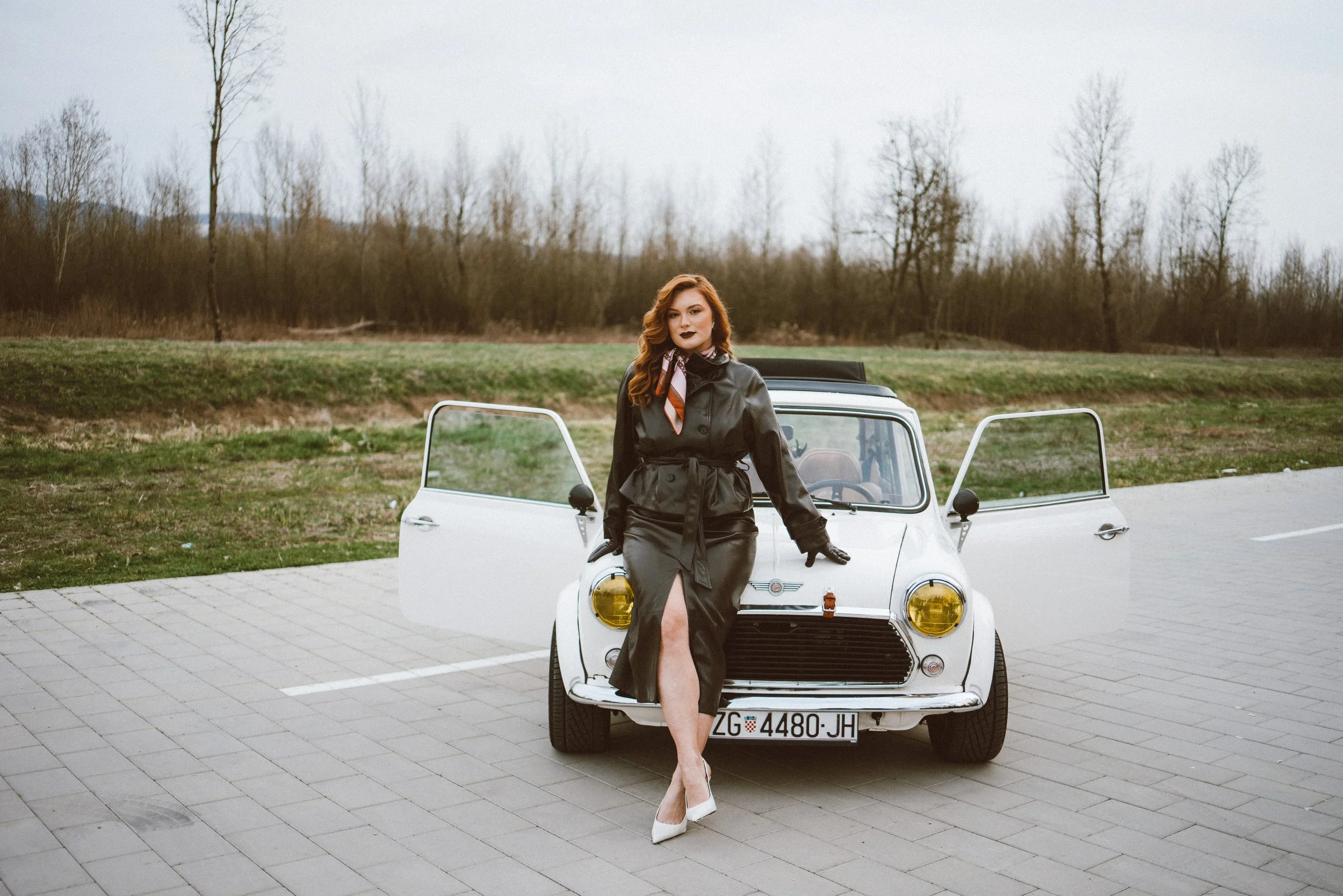 A woman with red hair dressed in a black trench coat, white high heels, and a colorful scarf, standing in front of a white vintage car with yellow headlights, on a paved area with grass and leafless trees in the background.