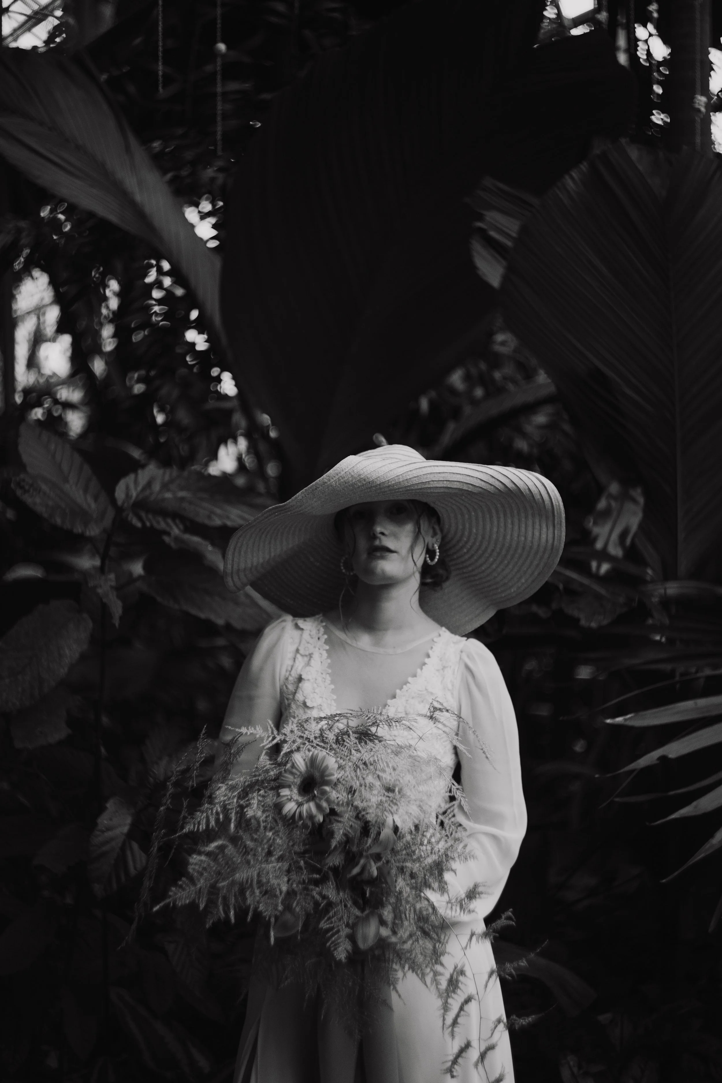 A woman in a large straw hat and a white dress holding a bouquet of flowers in front of dense foliage.
