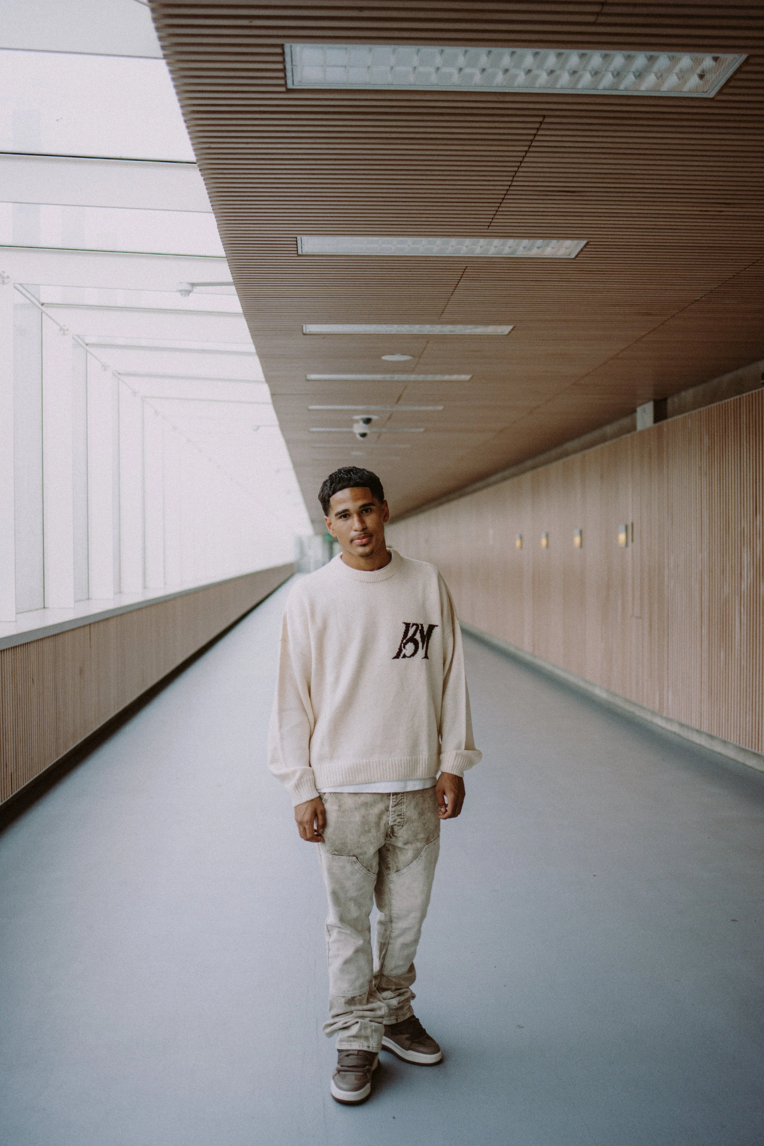 A young man stands in a modern corridor with wooden wall paneling and a ceiling with recessed lighting. He is wearing a cream-colored sweatshirt with black text, beige pants, and sneakers.