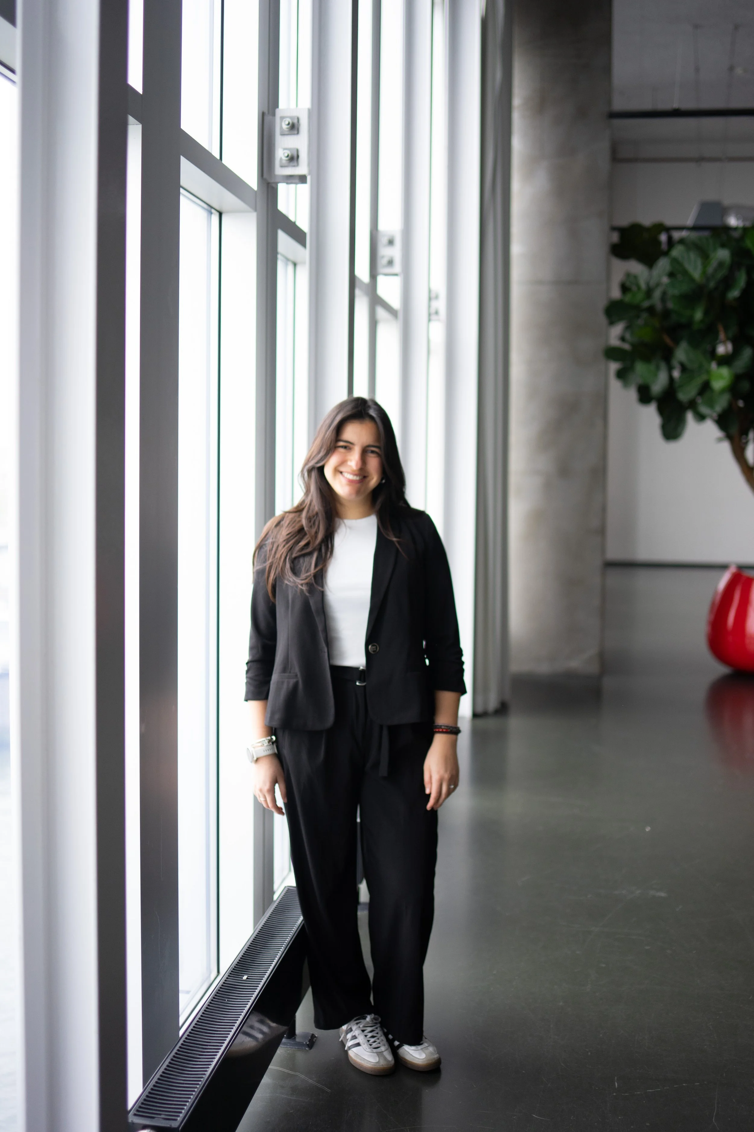 Smiling woman with long brown hair in a black suit and white sneakers standing next to a large glass window in a modern building.