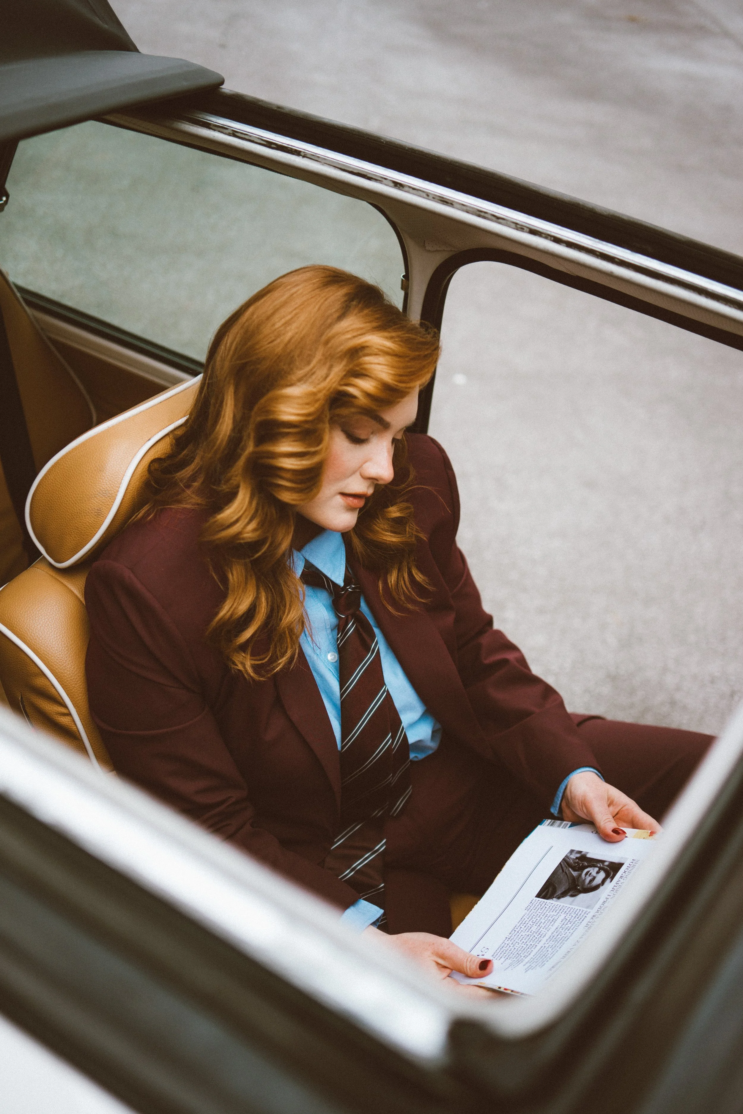 A woman with red hair sits in a car, reading a newspaper with a black and white photo of a person on the page.