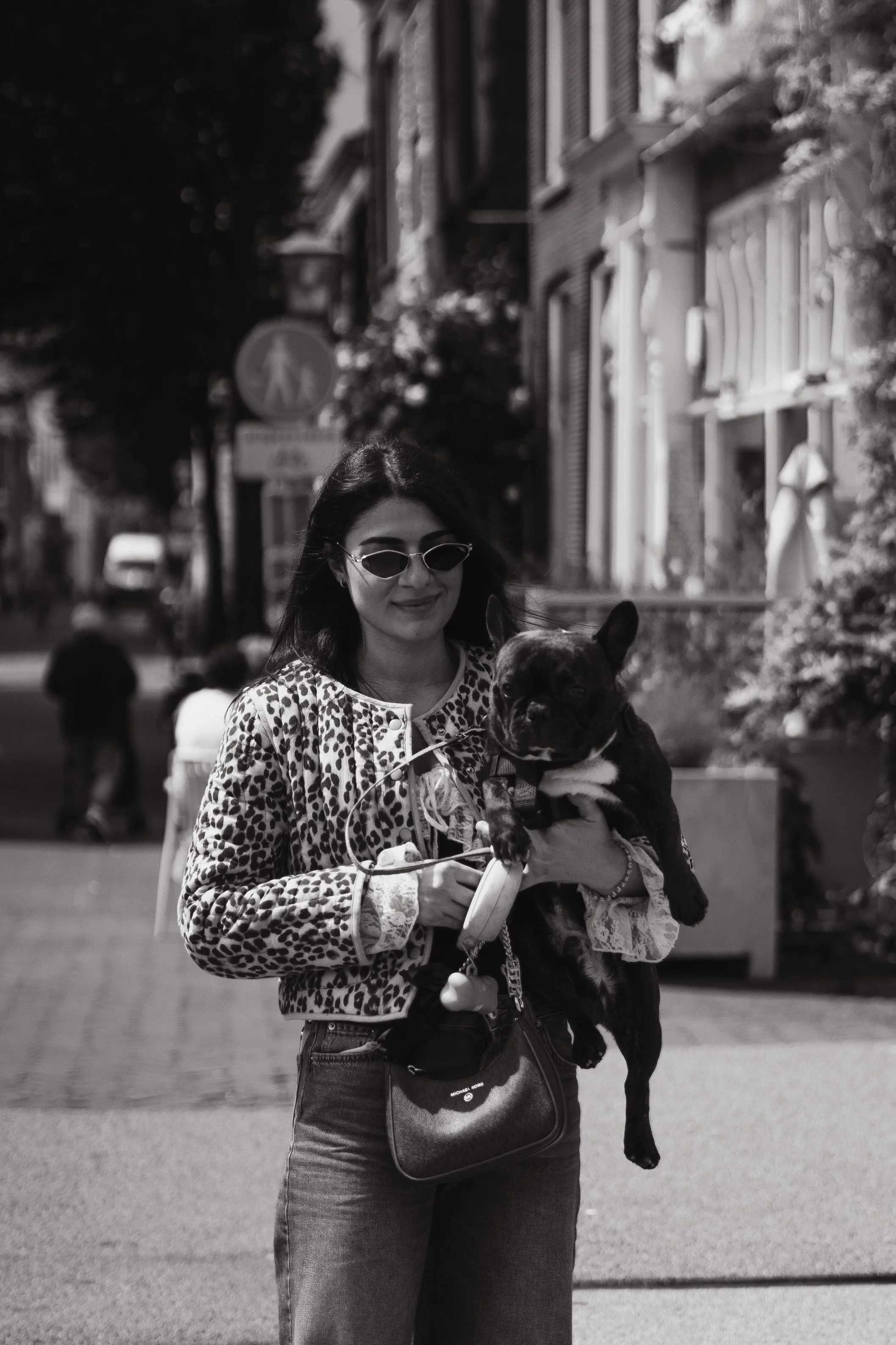A woman wearing sunglasses and a leopard print jacket is holding a small dog in her arms and a leash in her hand on a street with people in the background.
