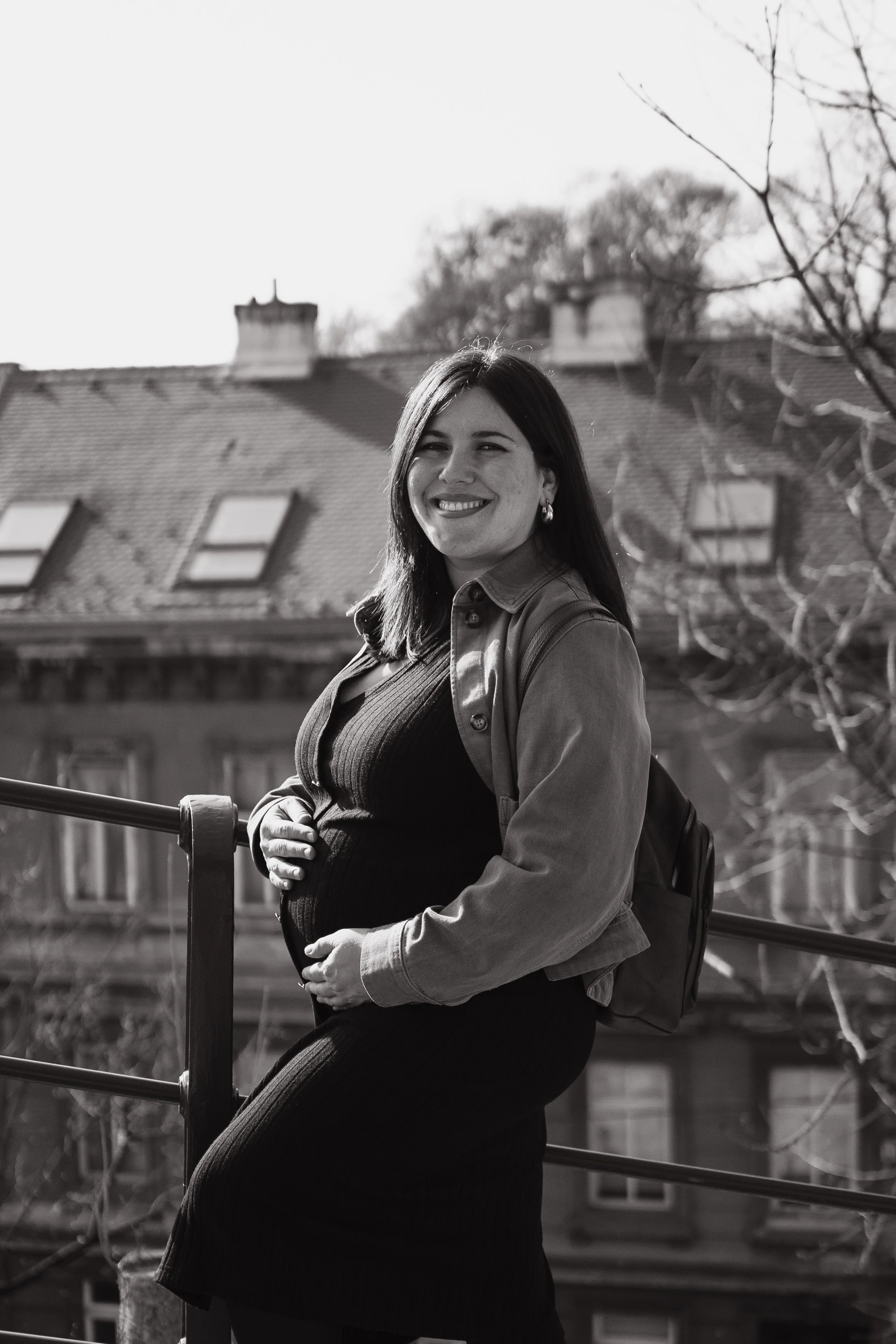 Black and white photo of a woman smiling and standing outdoors, with trees and rooftops in the background.