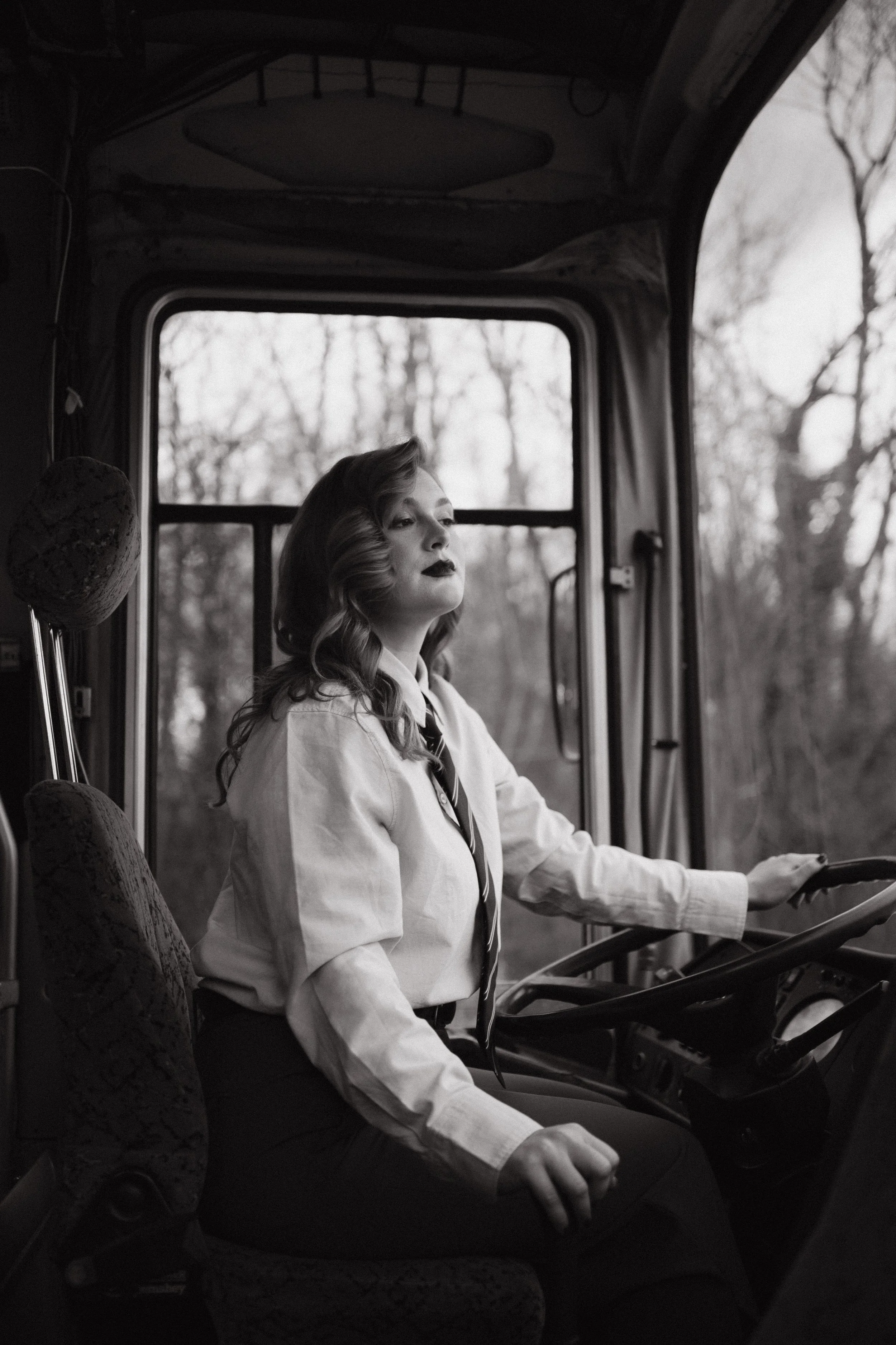 A woman with wavy hair and dark lipstick sitting in the driver's seat of a bus, with her left hand on the steering wheel, looking out the window. The background shows trees and a cloudy sky.