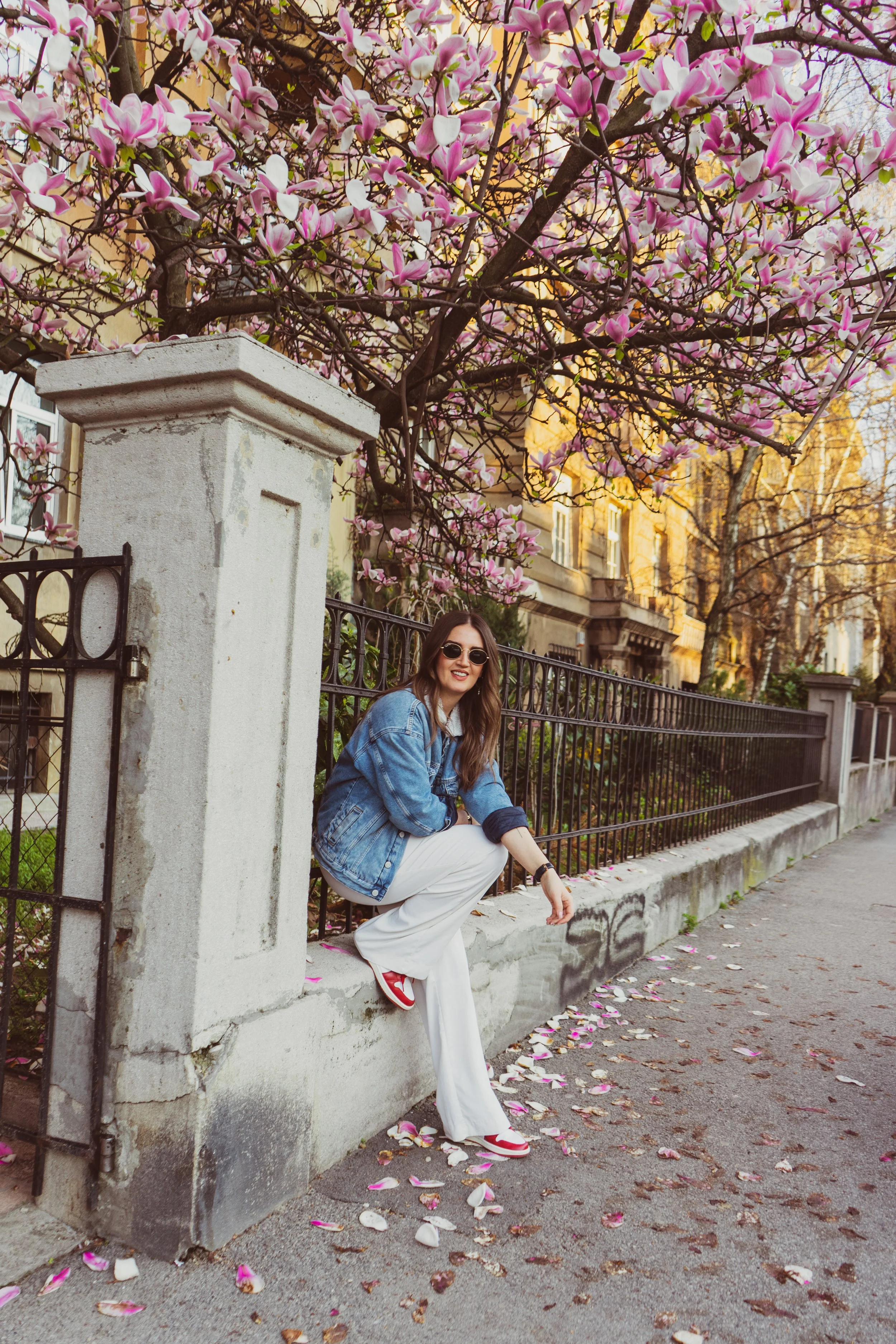 A woman with long brown hair wears sunglasses, a denim jacket, white pants, and red sneakers, sitting on a concrete ledge next to a black iron fence. She is smiling and squatting, with a tree with pink and white blossoms and building facades with yel