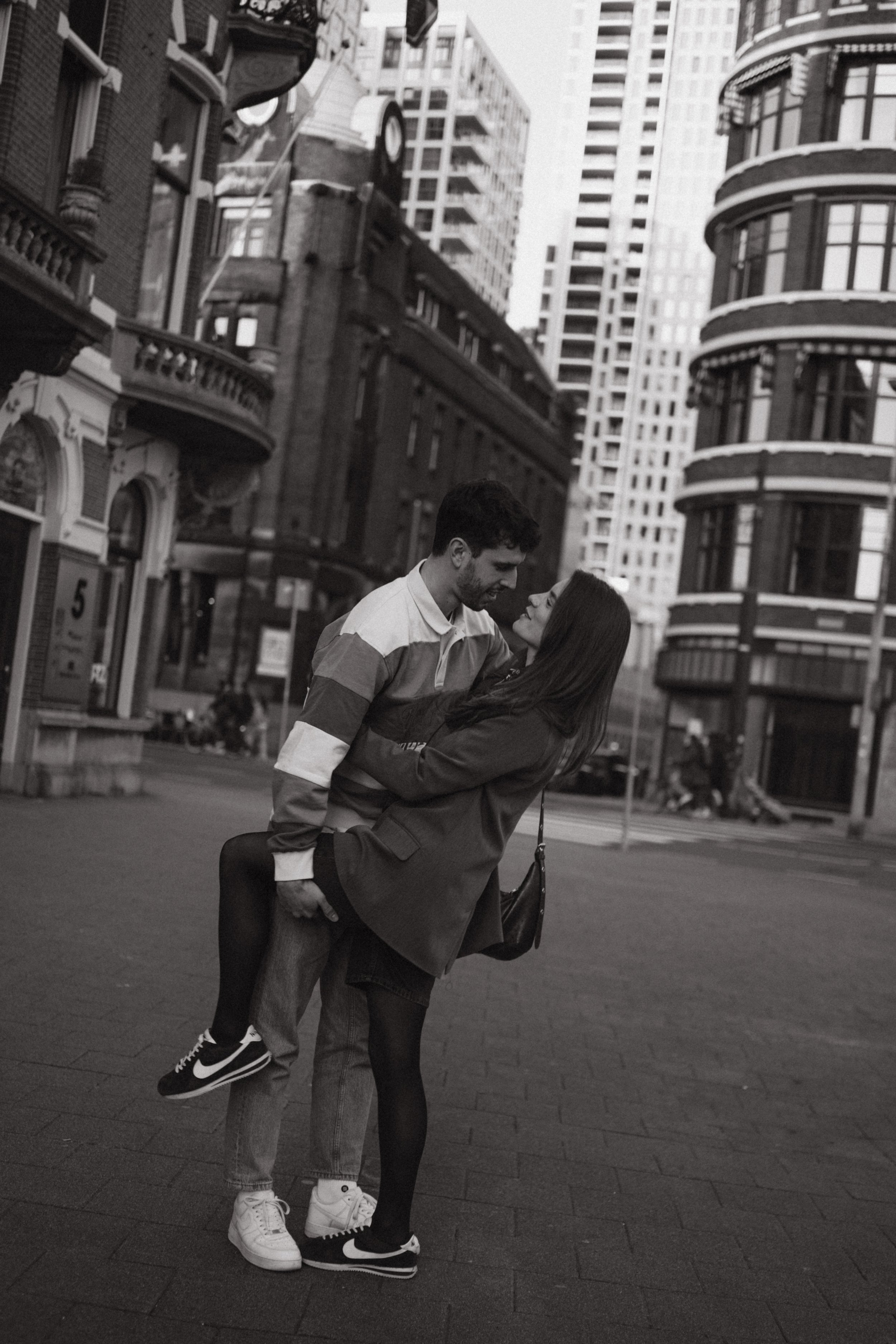 A black-and-white photo of a young couple in an urban setting, with tall buildings surrounding them. The man is lifting the woman, and they are looking at each other closely, smiling. The woman has long hair and is wearing a jacket and tights; the ma