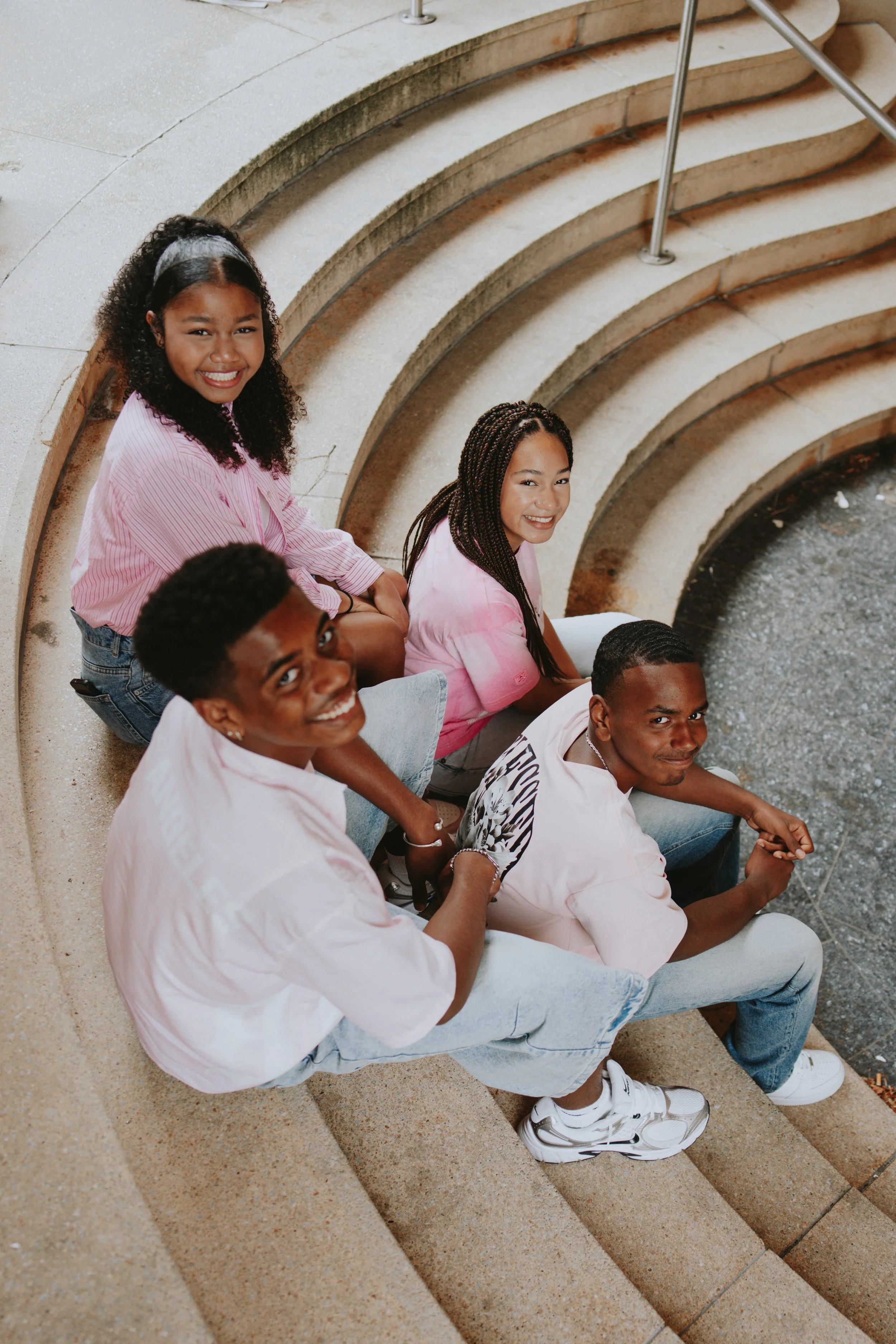 Group of five young people sitting and standing on outdoor concrete steps, smiling at the camera, wearing casual clothes.