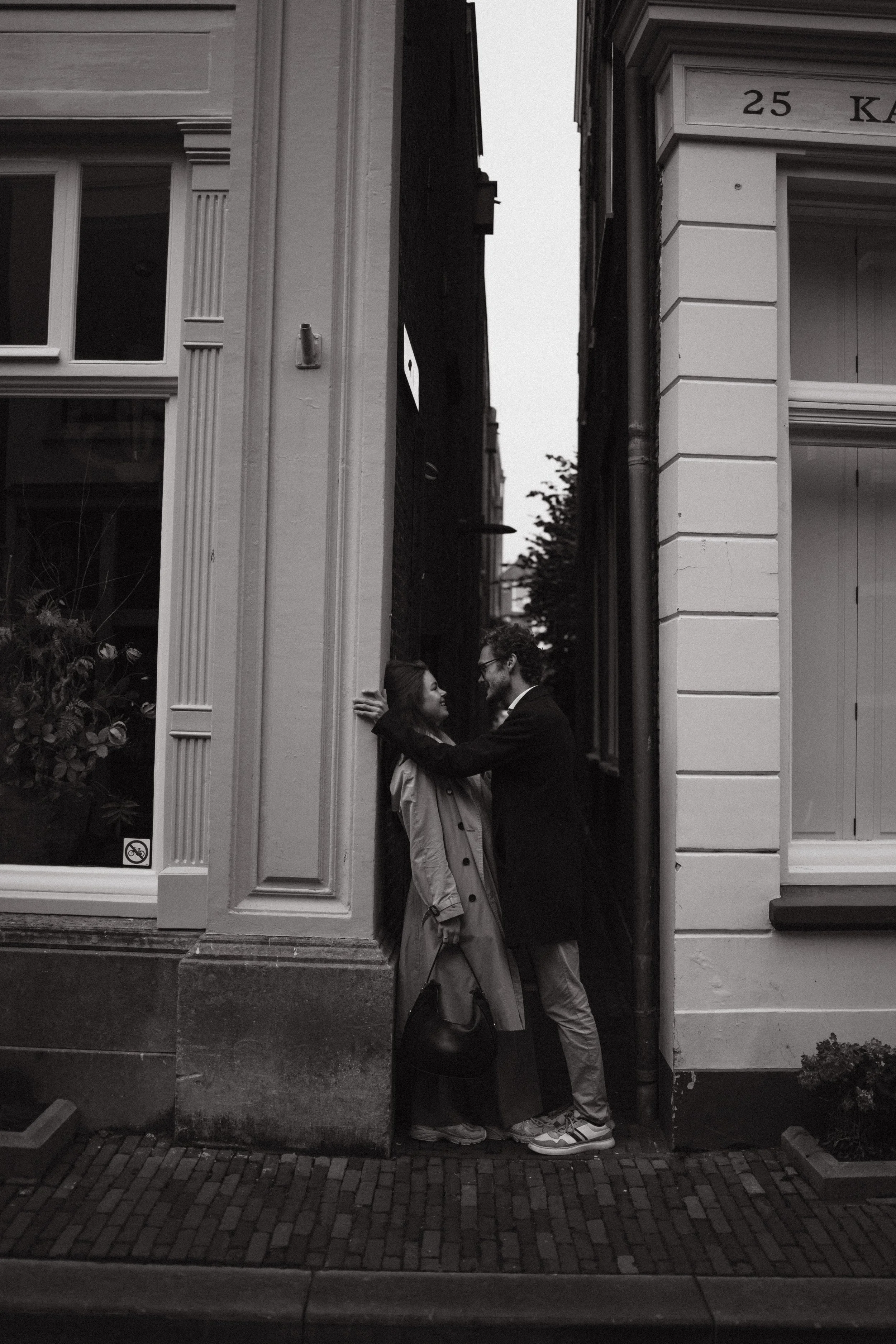 A black-and-white photo of a man and woman standing close and facing each other in a narrow alleyway between buildings. The woman is leaning against the wall, and the man is smiling and touching her face. They are close and seem to be sharing an inti