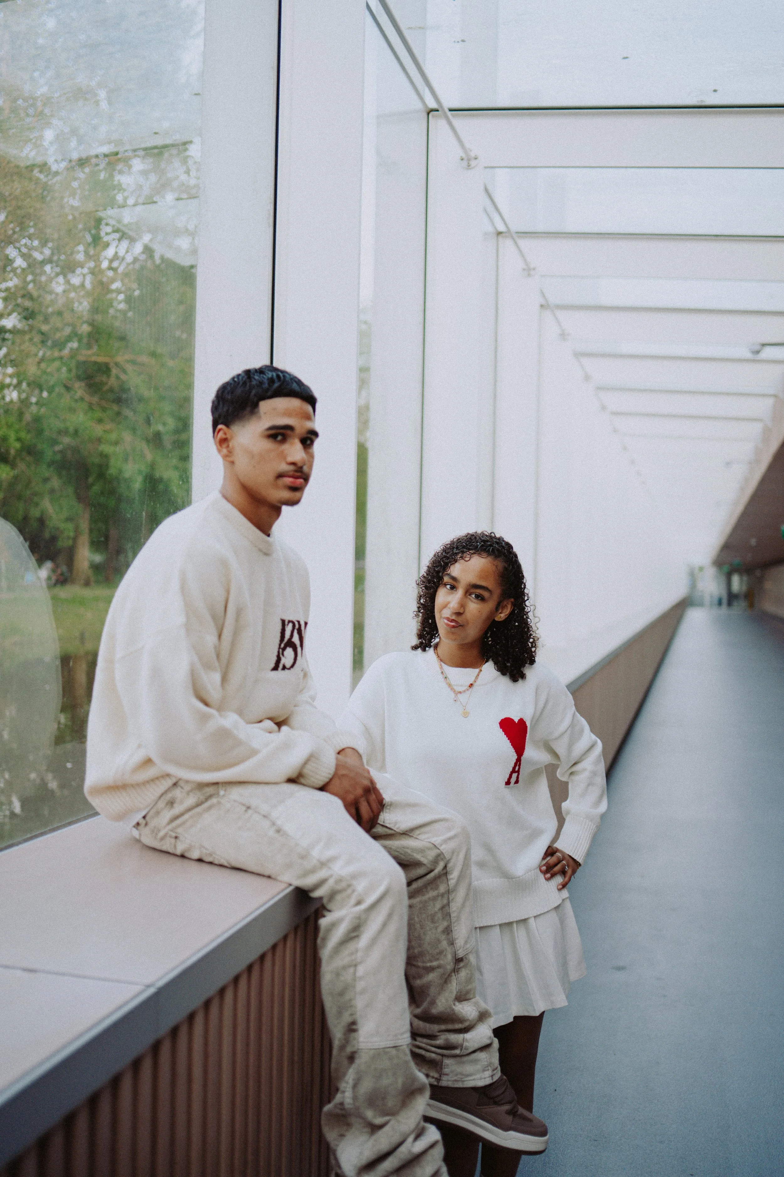 Two young adults, a man and a woman, sitting on a bench inside a modern building with large glass windows. The man, with short dark hair and a light cream-colored sweater, looks directly at the camera. The woman, with curly hair, wears a white sweate