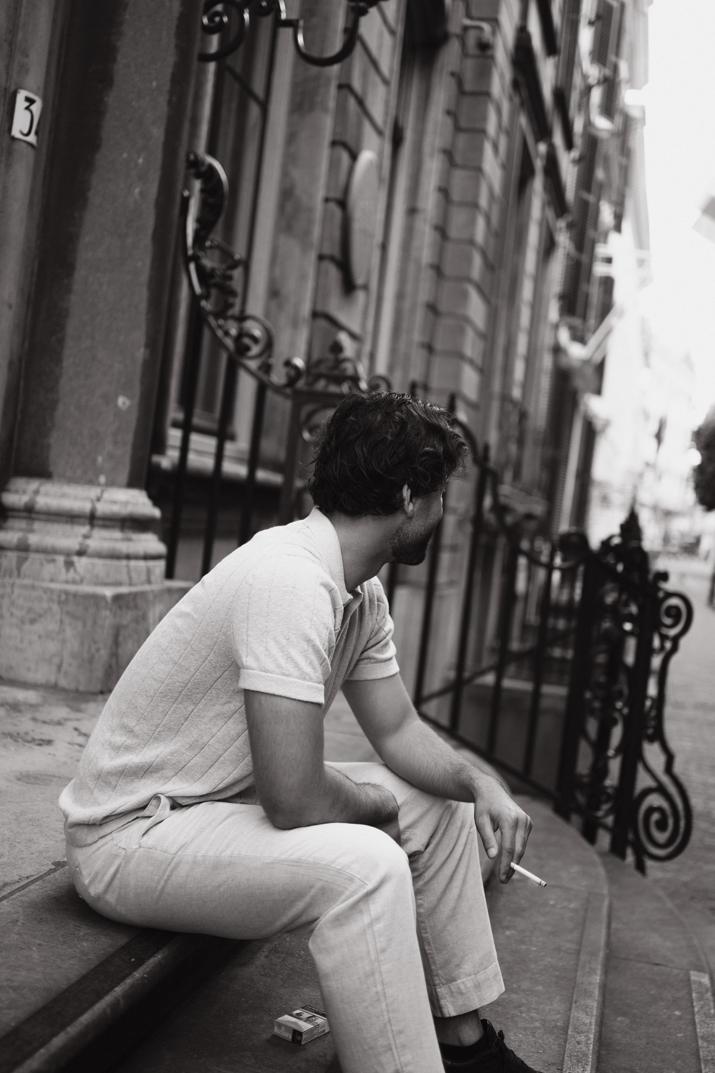 A man with curly hair and a beard sitting on a stone bench outside a building, holding a cigarette, wearing a light-colored short-sleeve shirt and pants. There is a pack of cigarettes on the ground near his feet, and a decorative wrought iron fence i