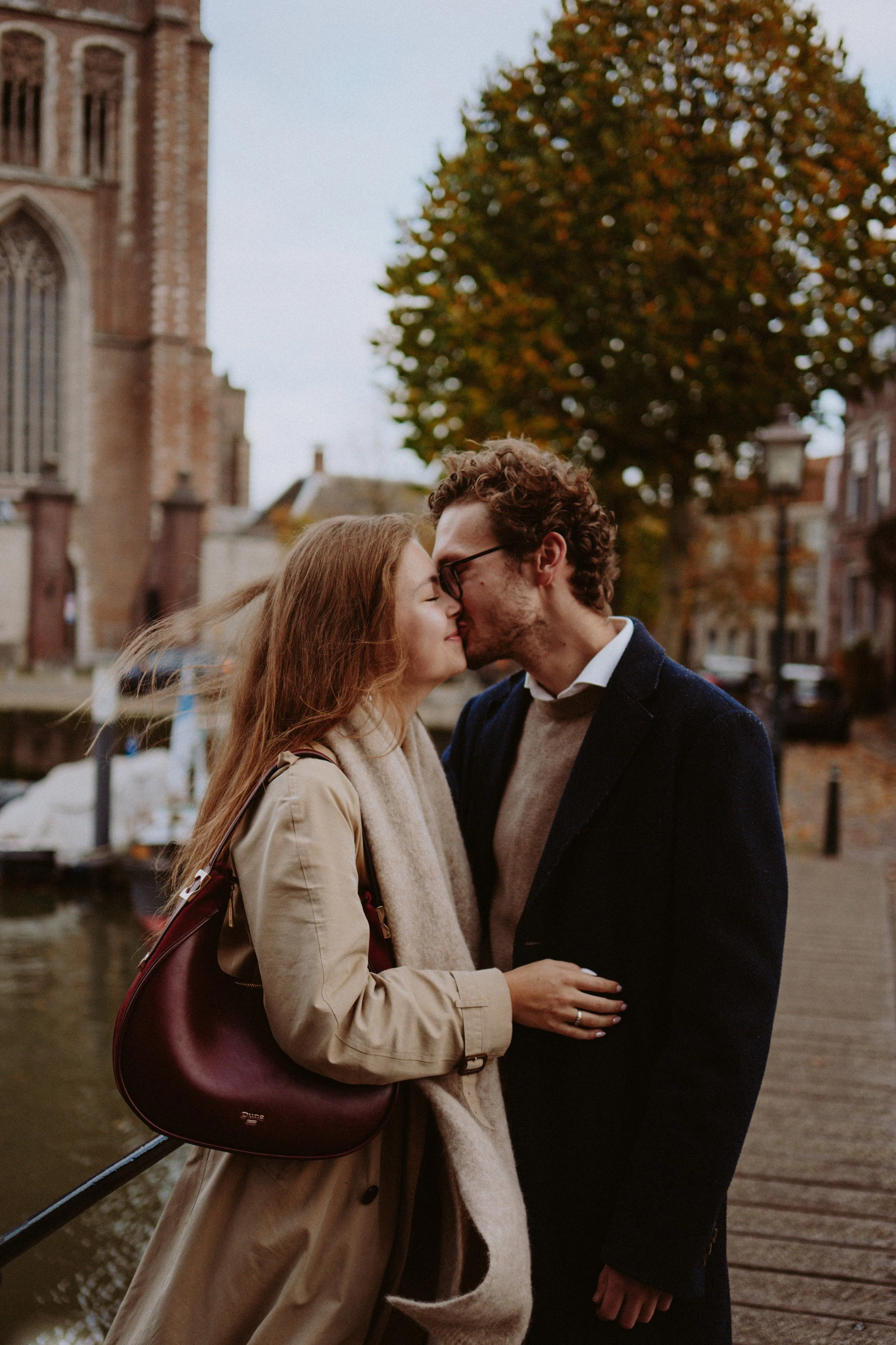 A couple is sharing a kiss on a bridge near a river with historic buildings and autumn trees in the background.
