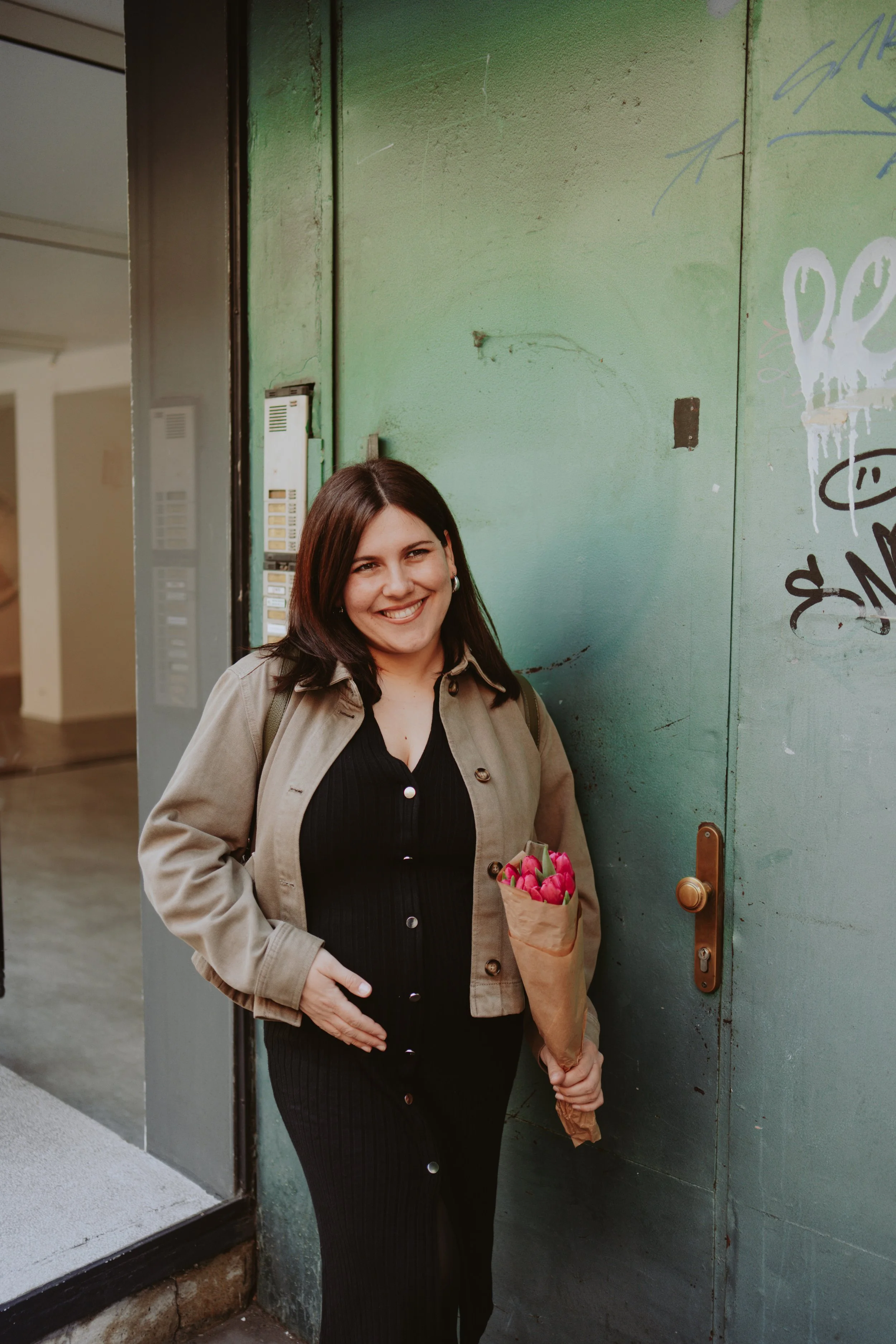 A woman with brown hair, wearing a beige jacket and black dress, smiling and holding a bouquet of pink tulips in an outdoor corridor near a green door with graffiti.